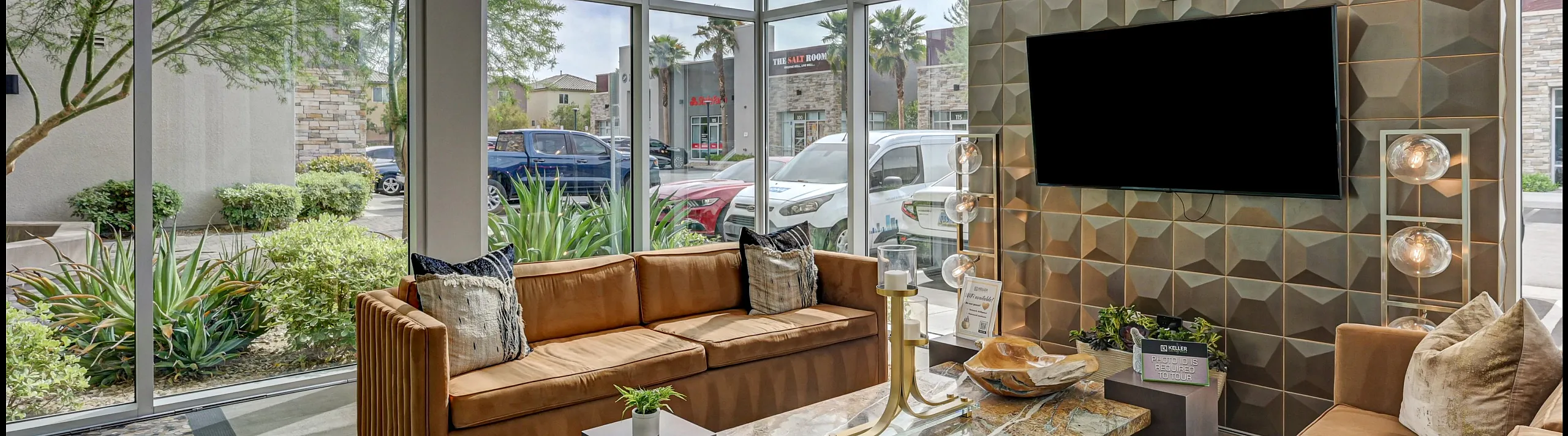 Modern apartment lounge with stylish brown sofas, geometric wall design, and floor-to-ceiling windows overlooking lush greenery.