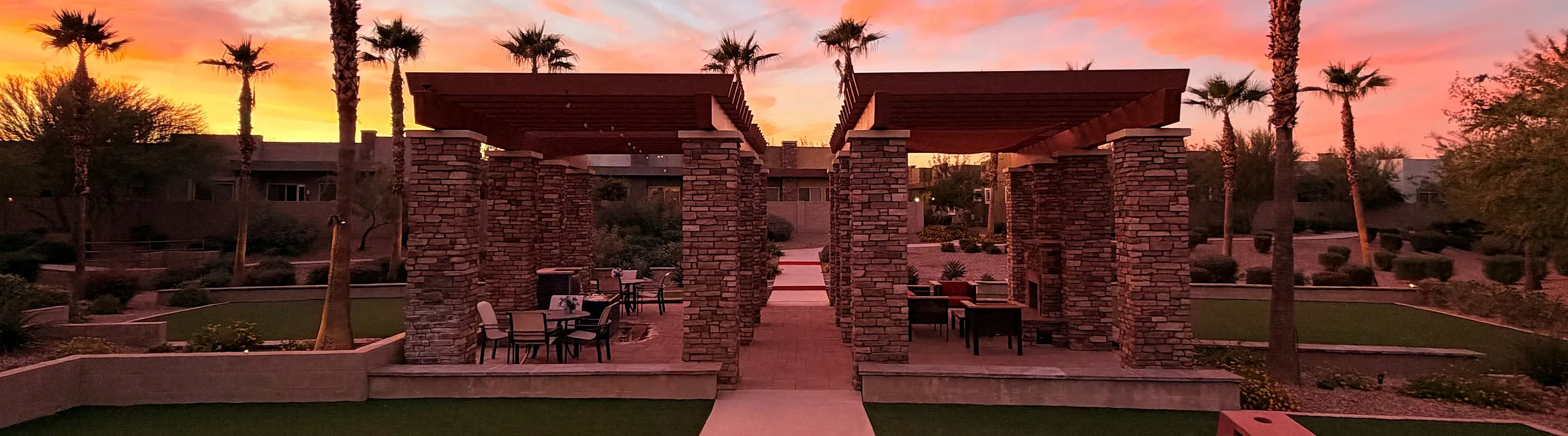 Enchanting courtyard with rustic stone pergolas and palm trees under a stunning pink and orange sunset sky.