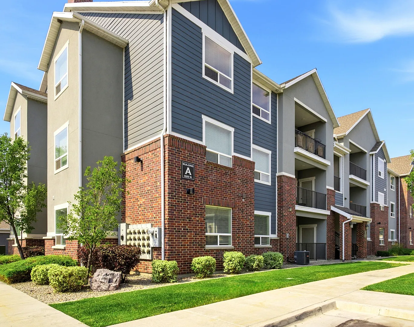 Charming modern apartment complex with brick and grey exterior, surrounded by lush greenery and vibrant landscaping under a clear blue sky.