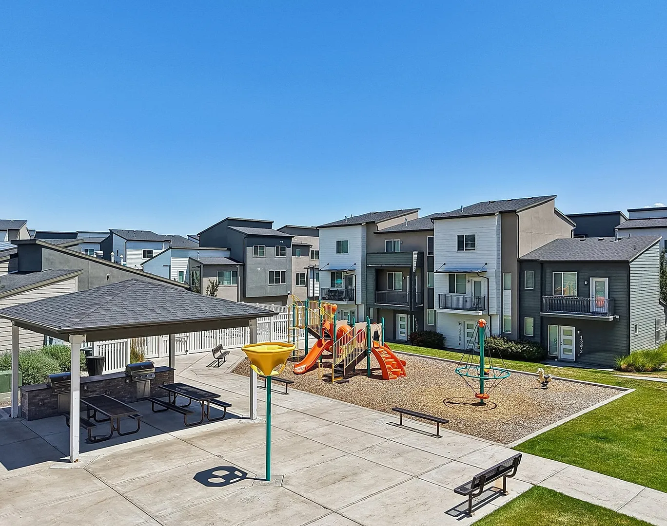 Modern apartment complex with vibrant playground and outdoor picnic area under a clear blue sky, perfect for family living.