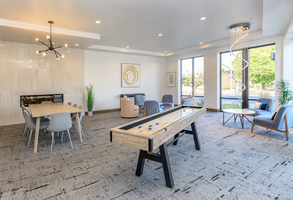 A modern, brightly-lit clubhouse/lobby space decorated with modern fixtures, a shuffleboard table, a large dining/conference table, and various lounge seating available.