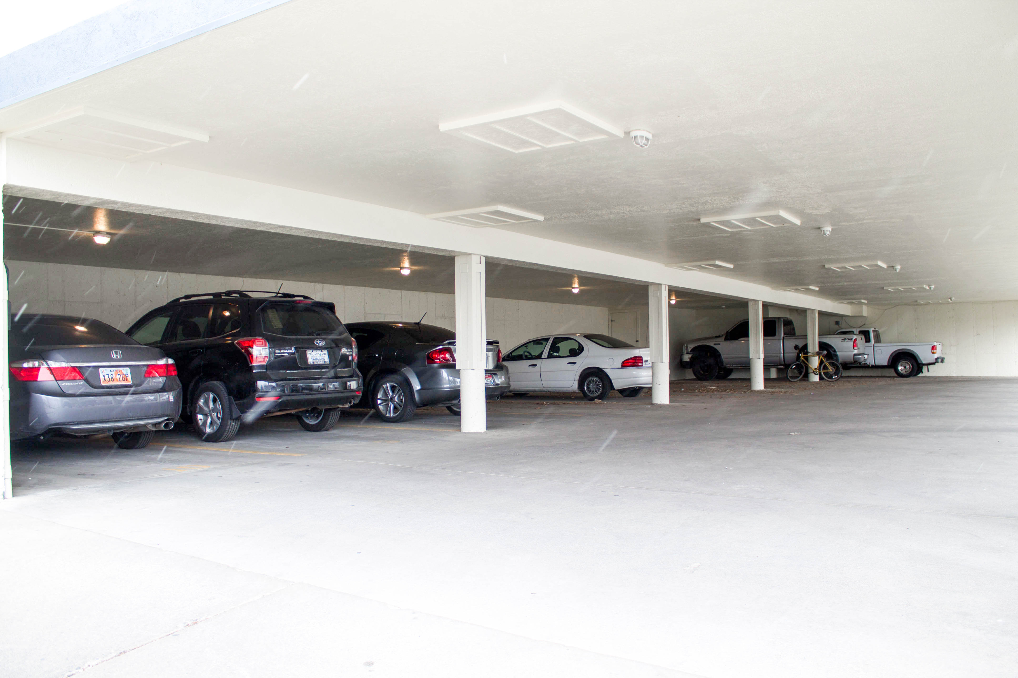 A covered parking garage with a low white ceiling and several white support pillars. Various cars and a silver pickup truck are parked in the spaces, and a yellow bicycle is leaned against one of the central pillars