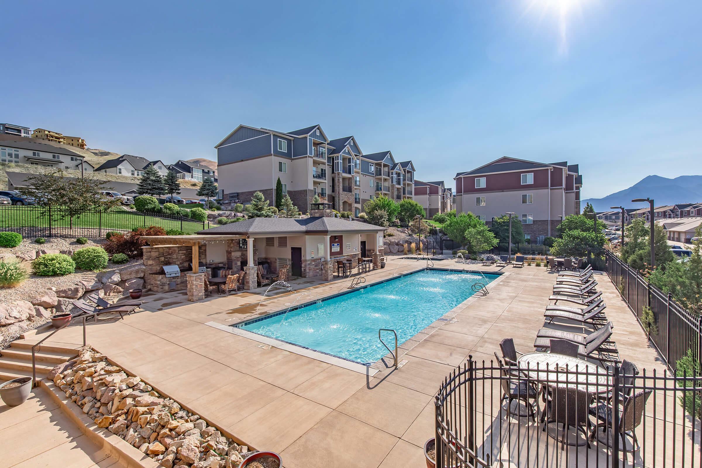 Charming apartment complex with a sunlit pool area, surrounded by lush landscaping and cozy outdoor seating, perfect for relaxation.
