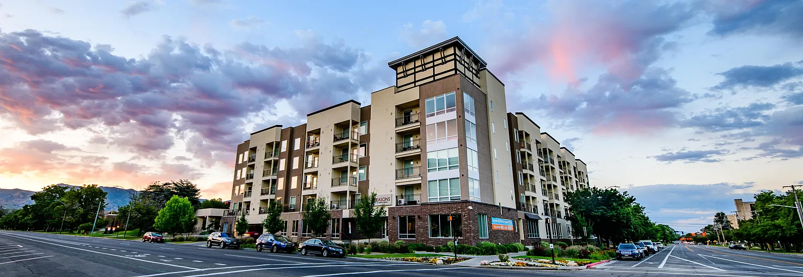 Modern apartment building with a stylish facade and lush surrounding greenery, set against a picturesque sunset sky.