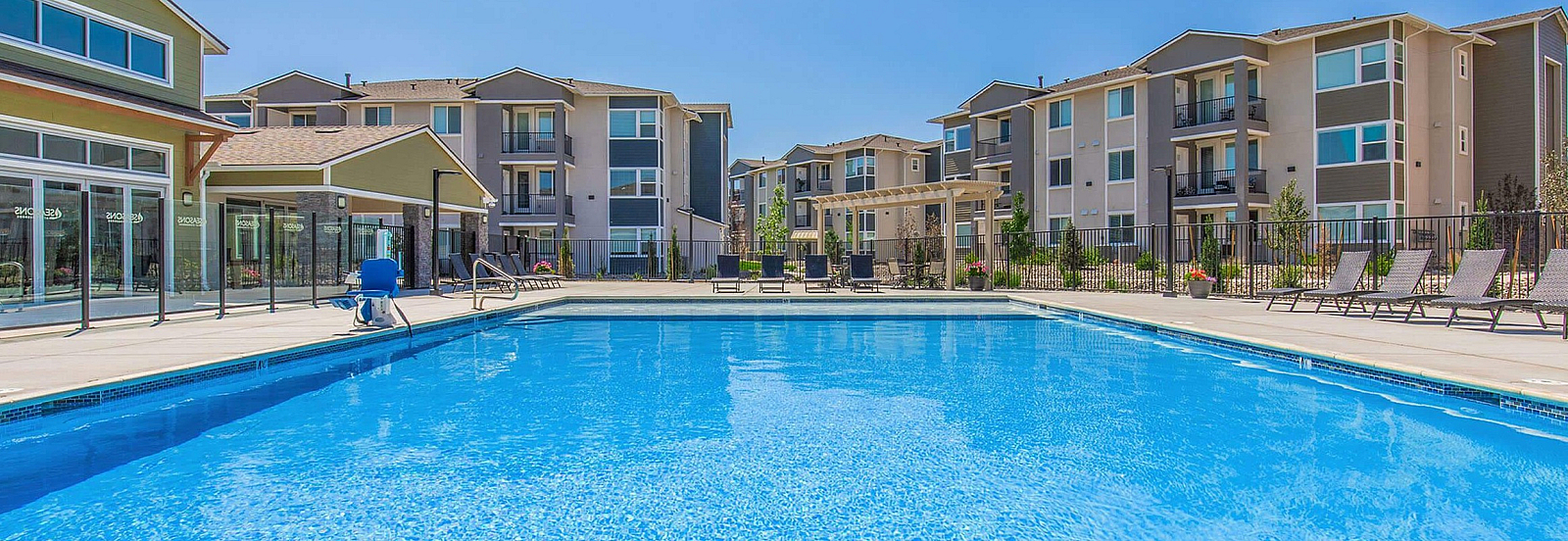 Modern apartment complex with stunning outdoor pool, surrounded by sun loungers and contemporary buildings under a clear blue sky.
