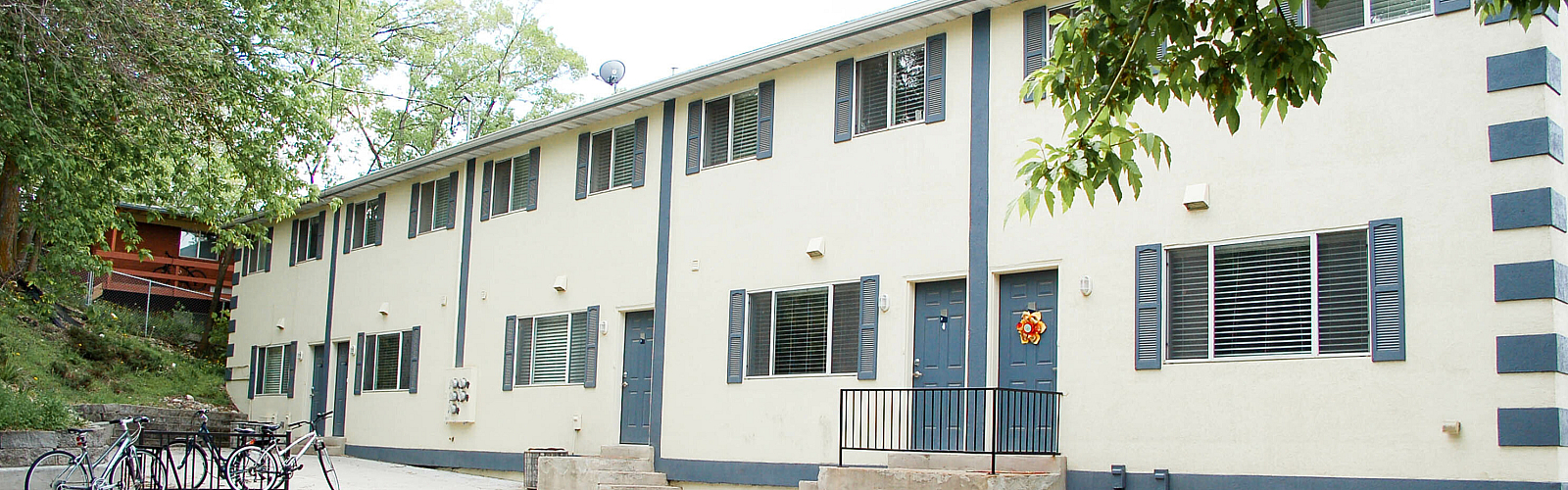 A two-story, multi-unit apartment building with cream-colored exterior walls and dark blue accents on the shutters, doors, and trim. Several concrete steps lead to the entrances, and a row of bicycles is parked on the left side under green