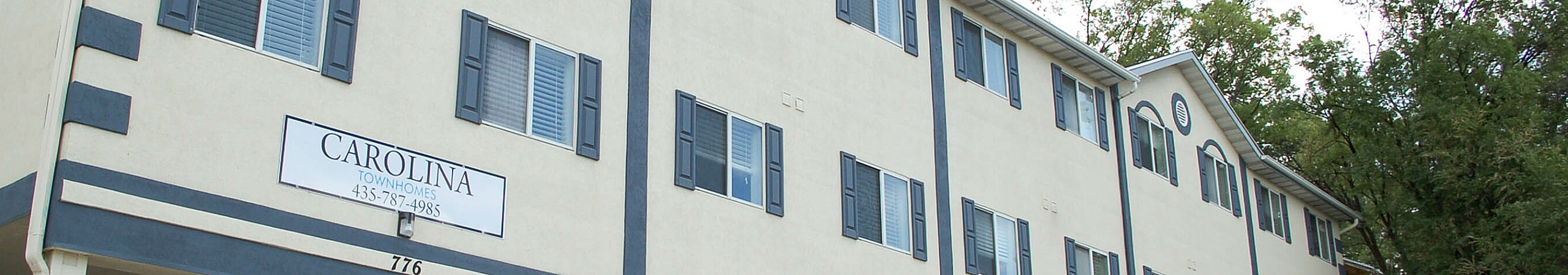 A wide, low-angle view of the cream-colored exterior of Carolina Townhomes. The building features dark blue shutters on the windows and a prominent white sign that reads "CAROLINA TOWNHOMES" above the phone number 435-787-4985.