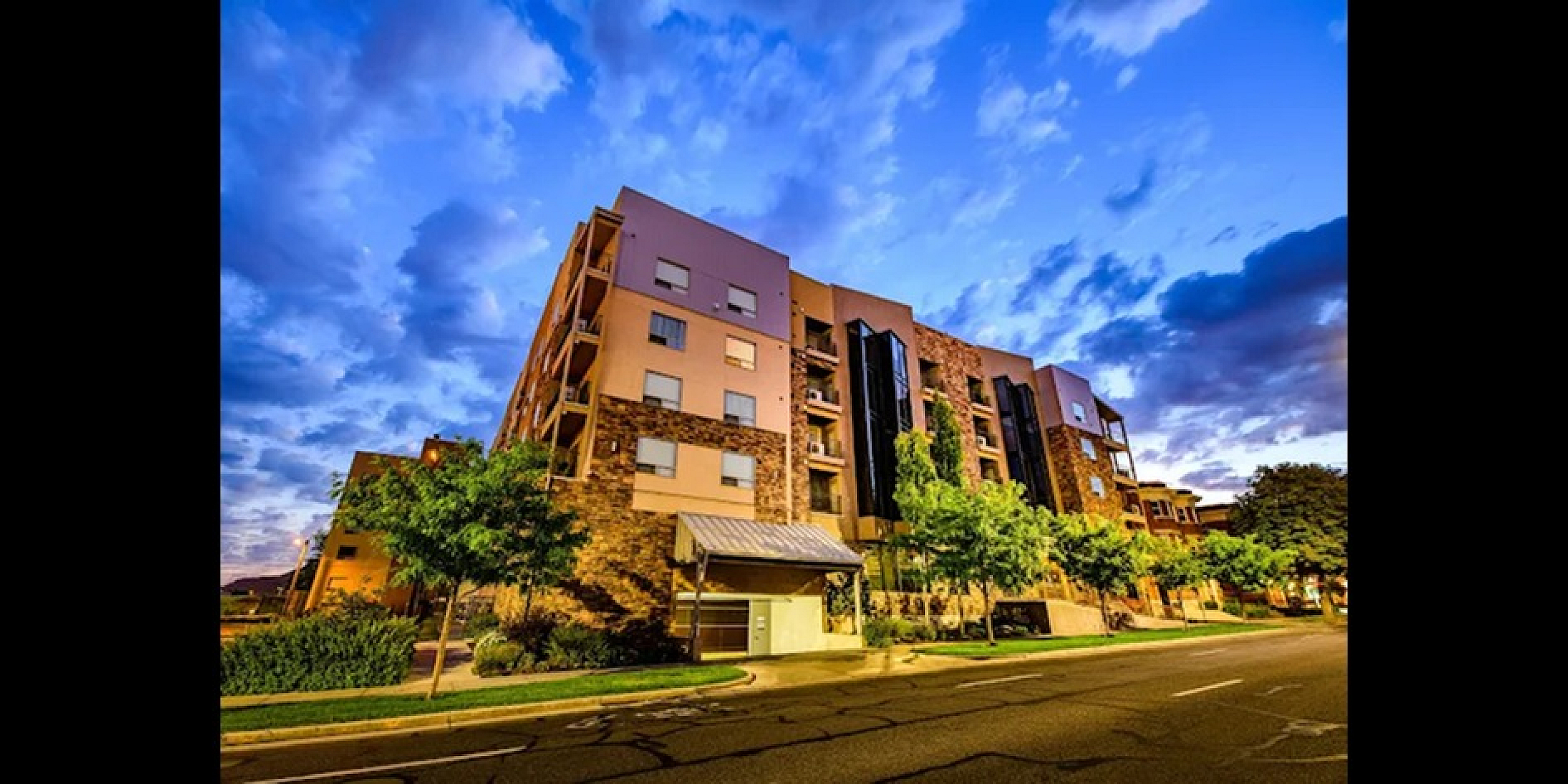 Elegant apartment building with modern architecture set against a vibrant sunset sky, surrounded by lush greenery on a calm street.