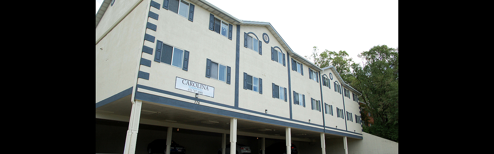 Exterior view of Carolina Townhomes, a three-story beige building with dark blue shutters and trim, featuring ground-level covered parking. A sign on the side of the building displays the property name, phone number (435-787-4985)