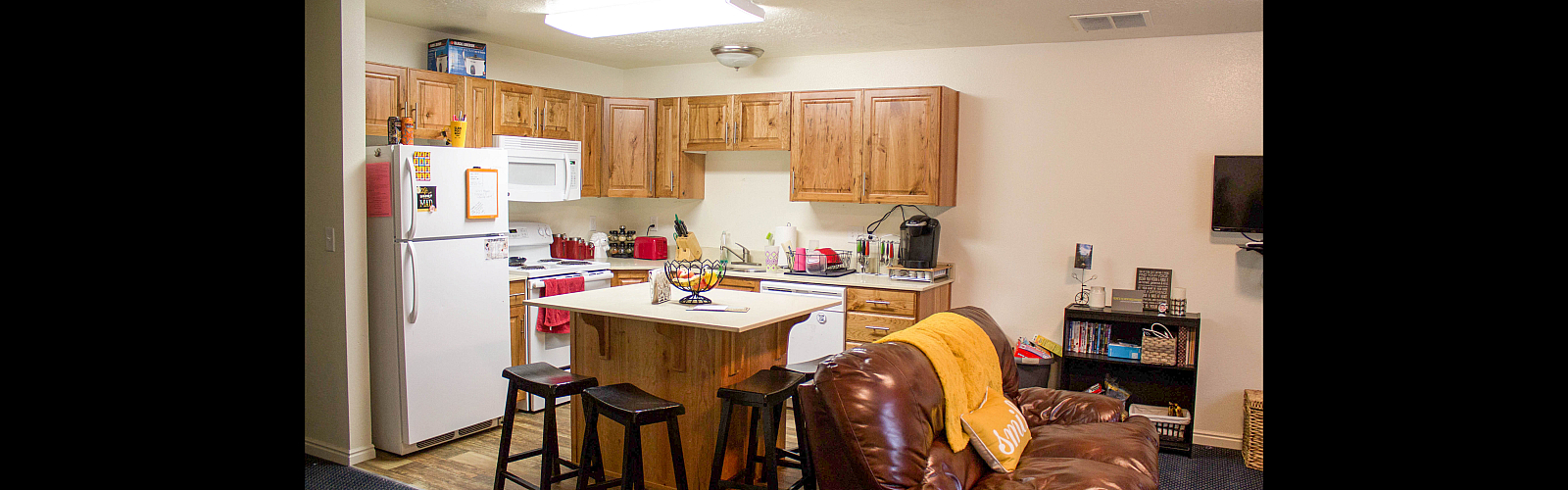 An open-concept kitchen and living area featuring light wood cabinets and white appliances. A small kitchen island with a white countertop is surrounded by three dark wooden bar stools. In the foreground, a brown leather sofa is draped with