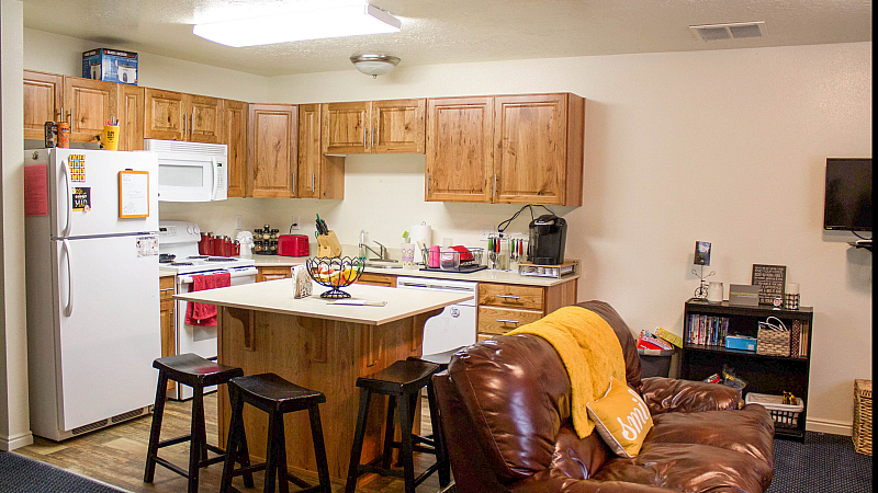 An open-concept kitchen and living area featuring light wood cabinets and white appliances. A small kitchen island with a white countertop is surrounded by three dark wooden bar stools. In the foreground, a brown leather sofa is draped with