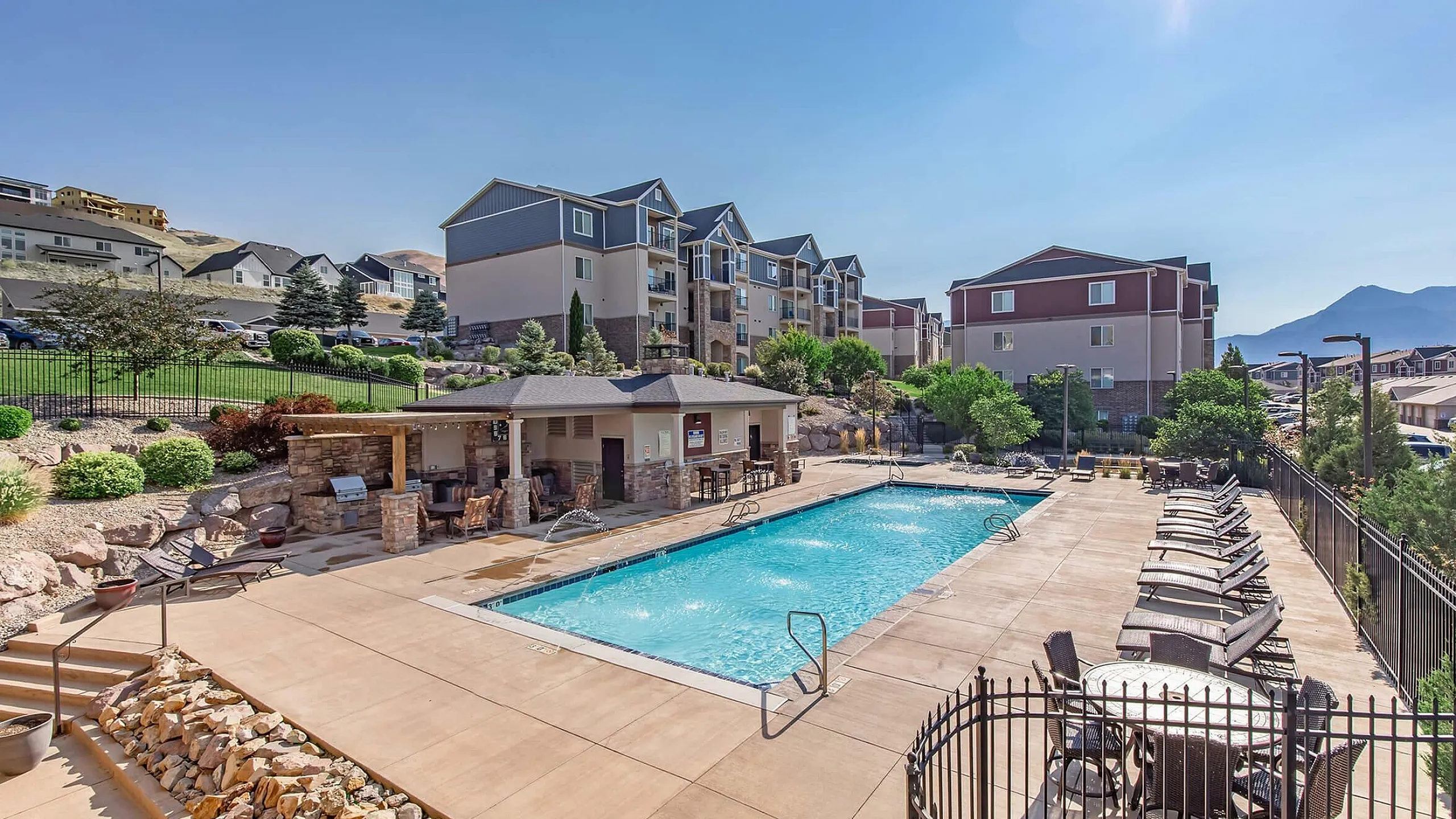 Inviting apartment complex with a sparkling outdoor pool, sun loungers, and mountain views, perfect for leisurely relaxation.