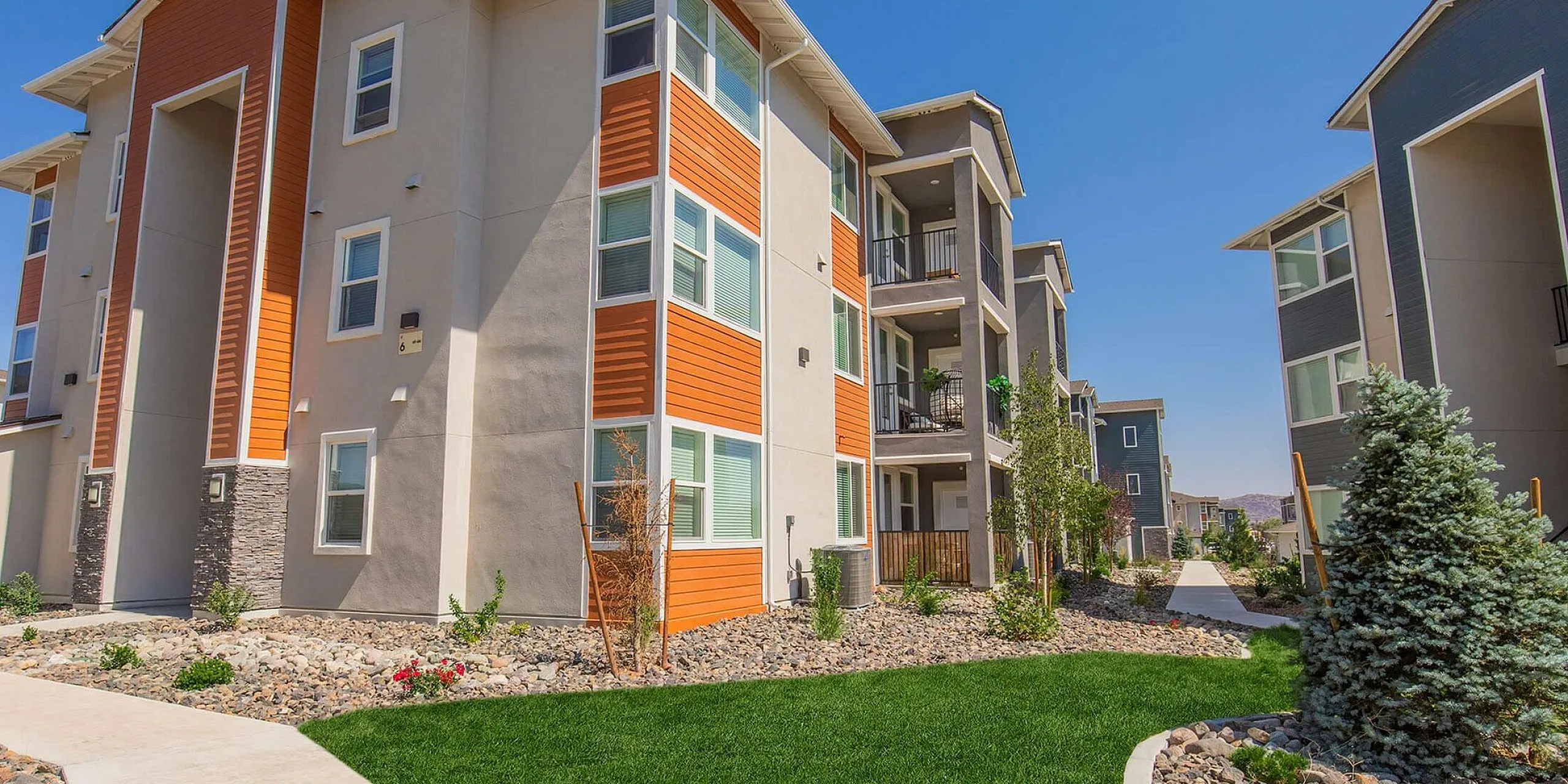 Modern apartment complex with vibrant orange accents, lush landscaping, and inviting pathways under a clear blue sky.
