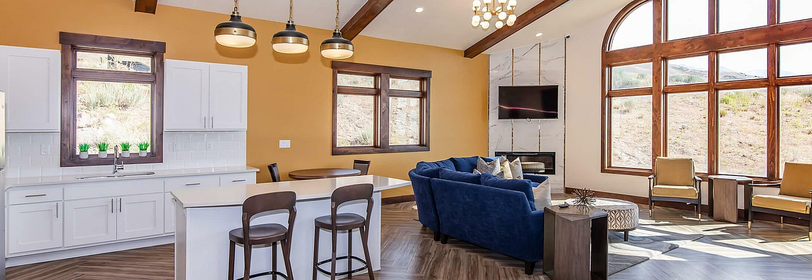 Bright open-concept living space with vaulted ceilings, elegant wood beams, and large arched windows. Features modern kitchen with white cabinetry and a cozy seating area with plush blue sofa and a contemporary fireplace.
