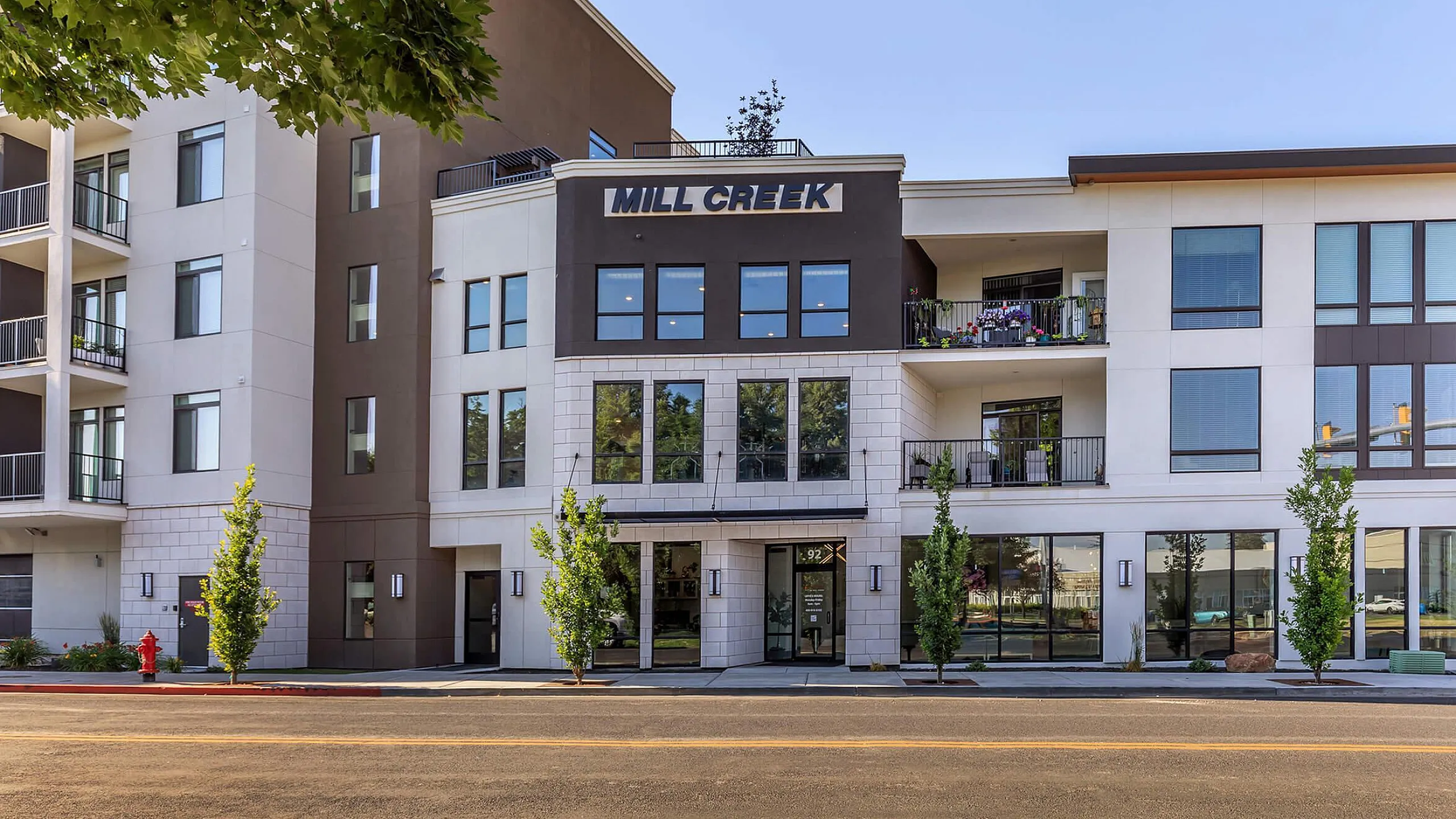 Front exterior of Mill Creek multi-family residential building displaying a mix of white stucco, dark brown siding, and light stone cladding. The building features large windows, private upper-floor balconies with seating and plants, street