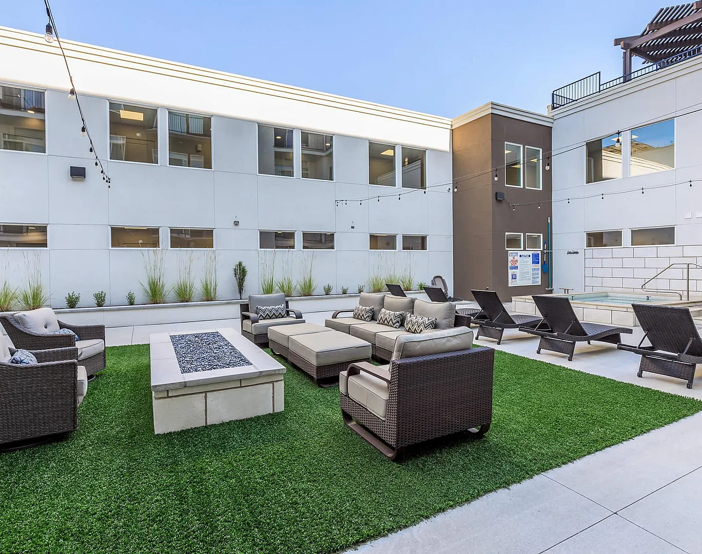 Modern apartment courtyard with outdoor lounge chairs, artificial turf lawn, built-in fire pit table, overhead bistro lights, and spa hot tub.