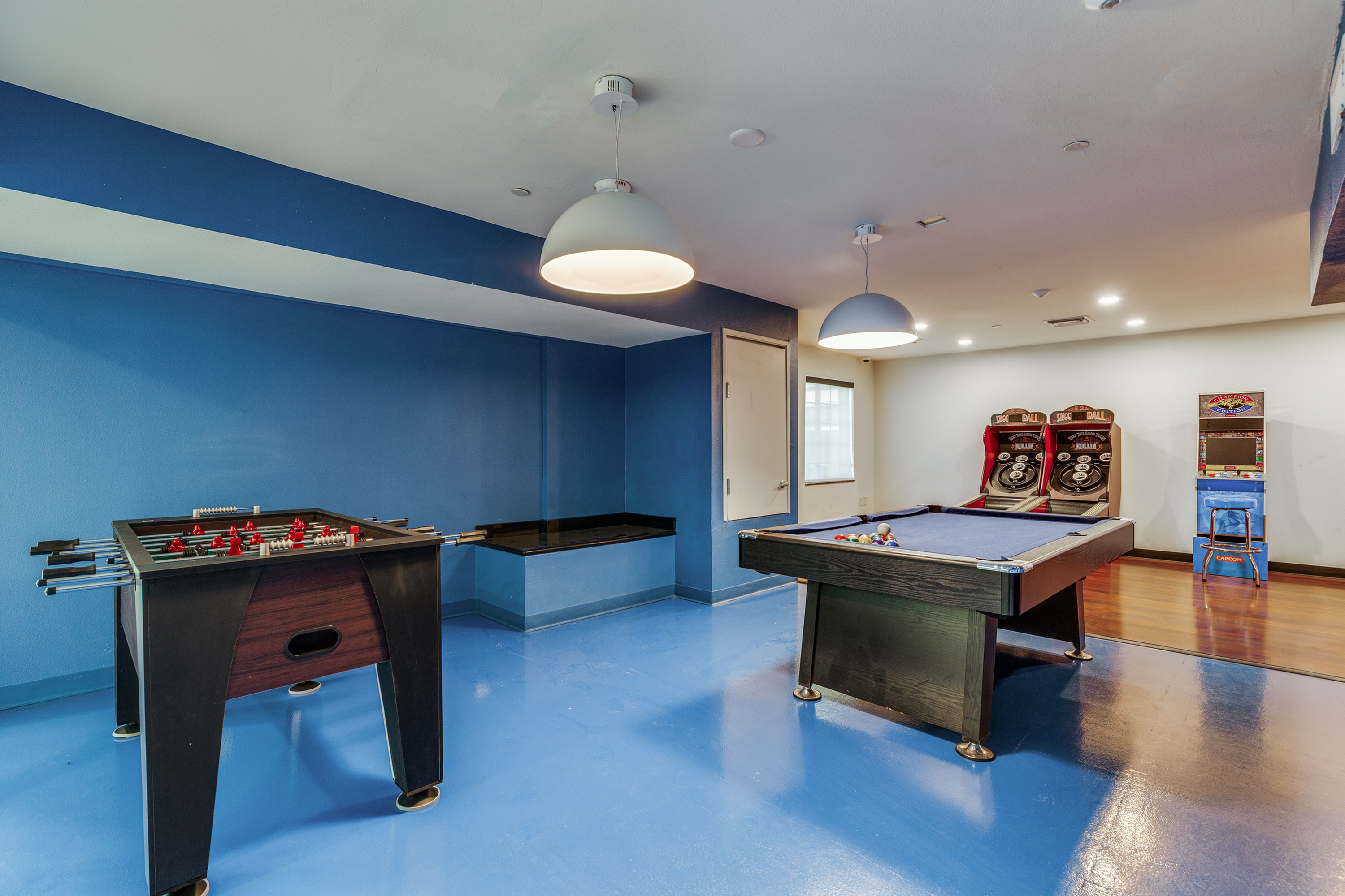 Vibrant game room with a sleek pool table, foosball table, and arcade games, featuring stylish blue decor and modern lighting.