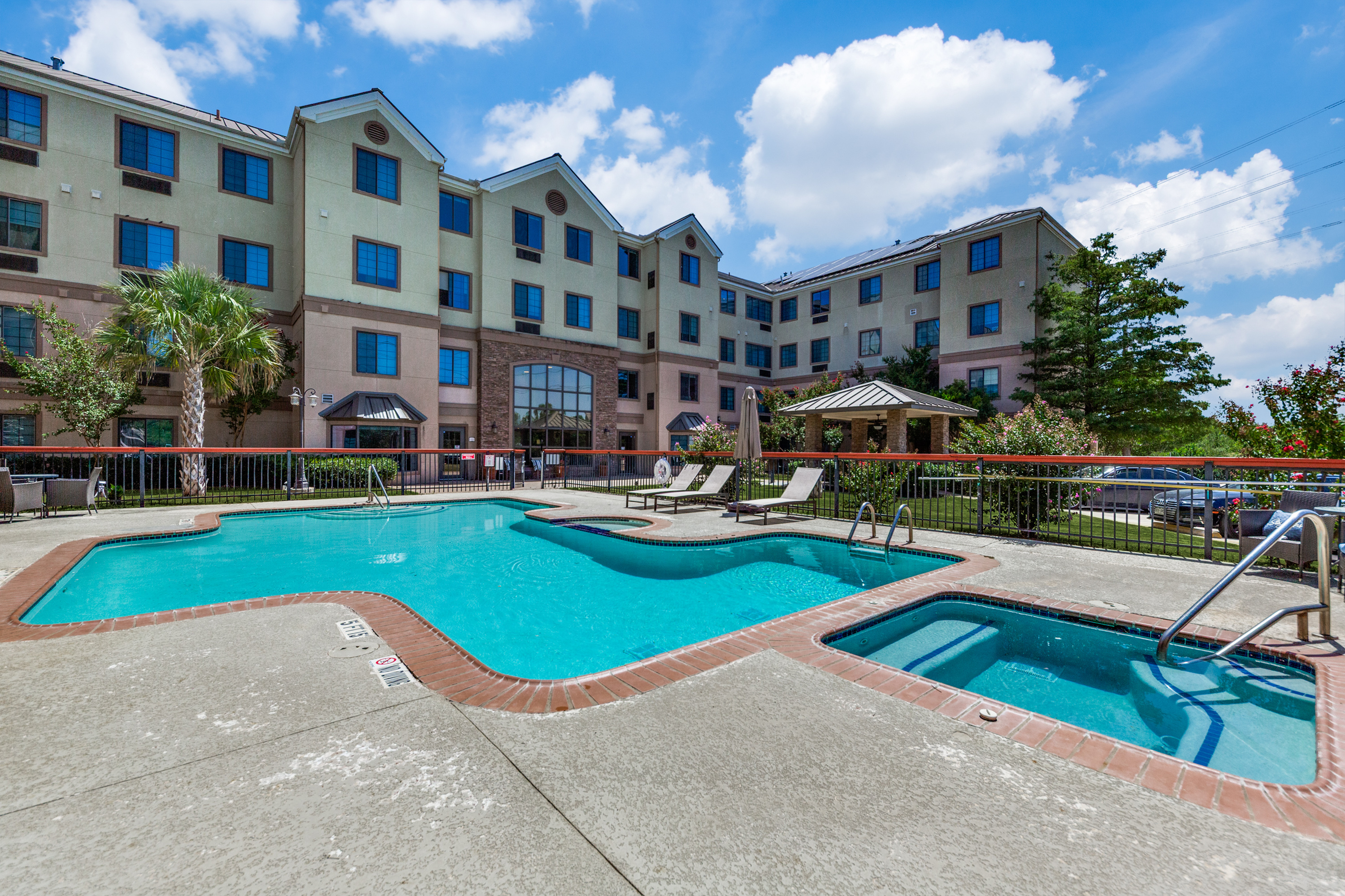 Inviting outdoor pool area with modern lounge chairs and beautiful landscaping, set against a stylish multi-story apartment building under a clear blue sky.