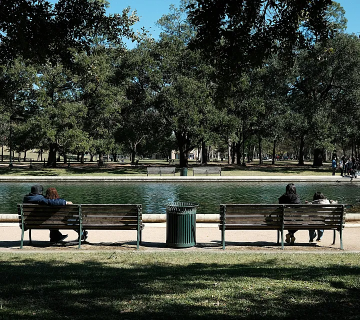 Peaceful park setting with two benches facing a tranquil pond, surrounded by lush green trees and visitors enjoying the serene environment.