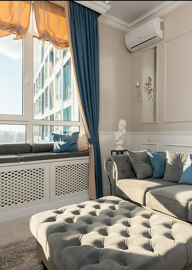 Charming living room with elegant tufted sofa, stylish ottoman, and large sunlit windows adorned with luxurious curtains.