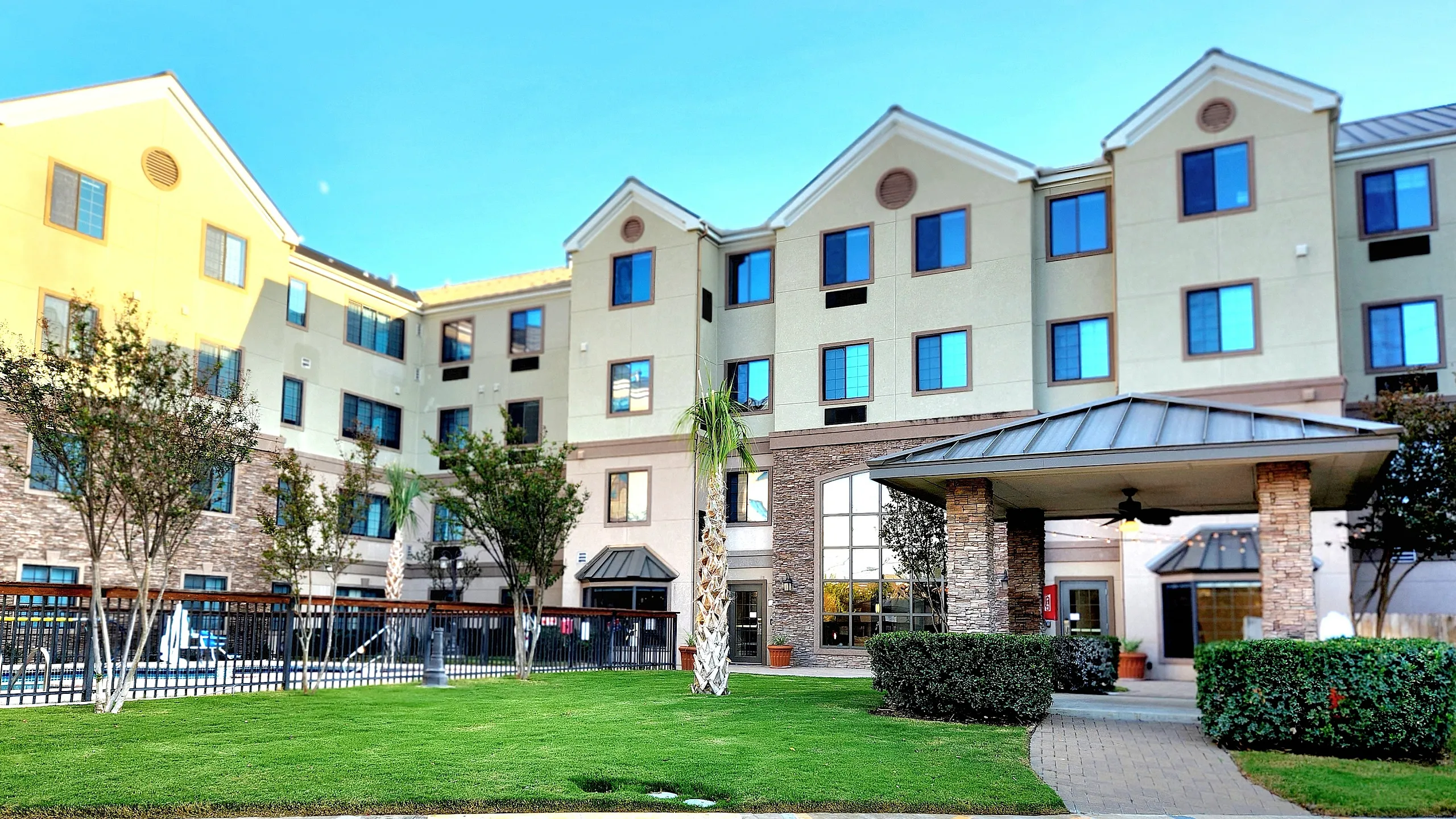 Charming apartment complex exterior featuring lush green lawns, a welcoming entrance, and vibrant landscaping under a clear blue sky.