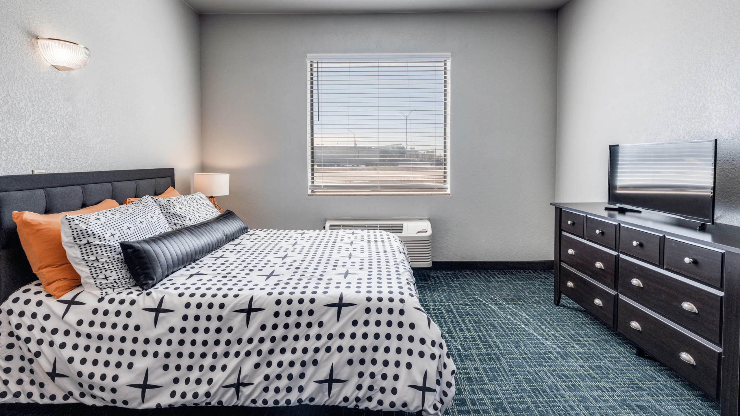 Chic bedroom with modern decor featuring a plush bed with stylish bedding, a sleek dresser with a flat-screen TV, and ample natural light through a large window.