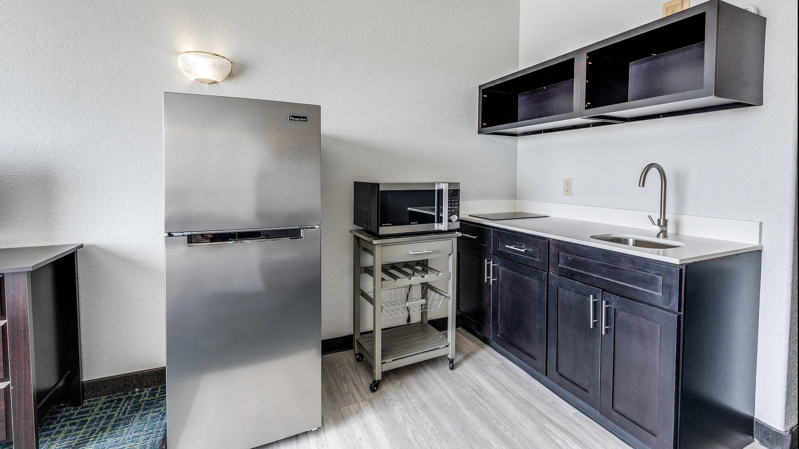 Modern kitchenette with sleek stainless steel fridge, elegant dark cabinets, and convenient microwave cart.