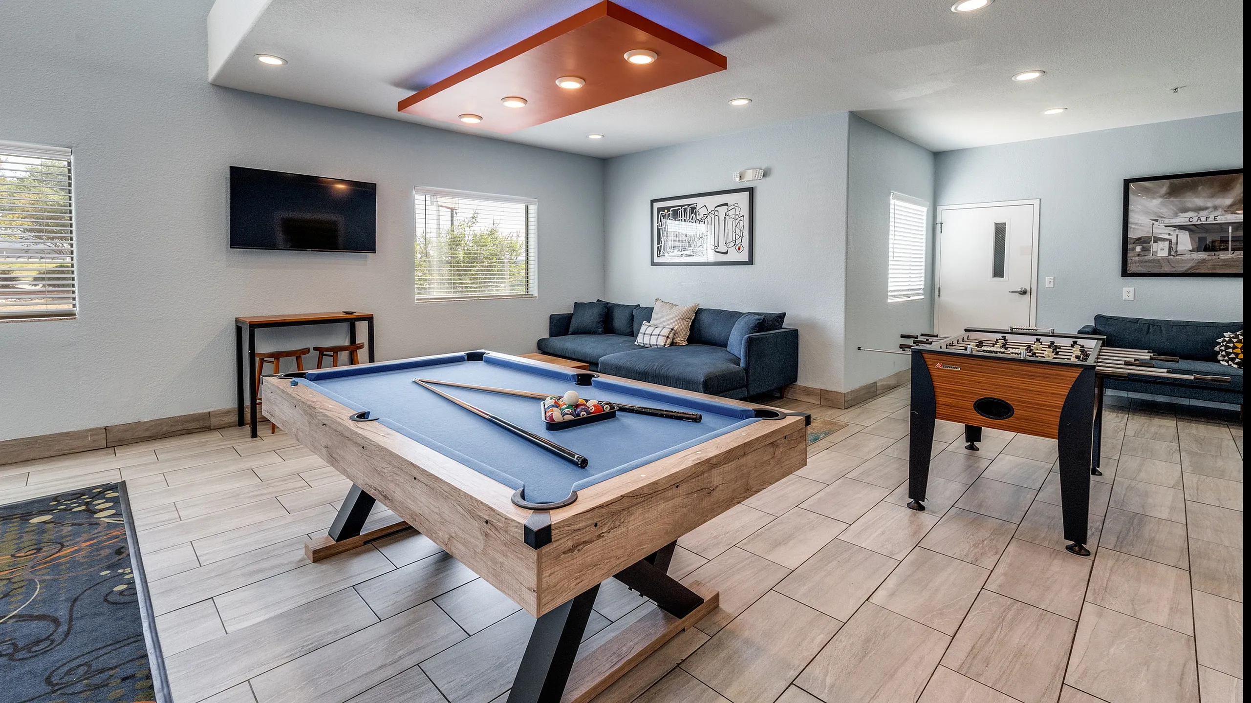 Inviting recreation room featuring a sleek pool table, comfortable blue sofa, wall-mounted TV, and a vibrant foosball table, perfect for entertainment.