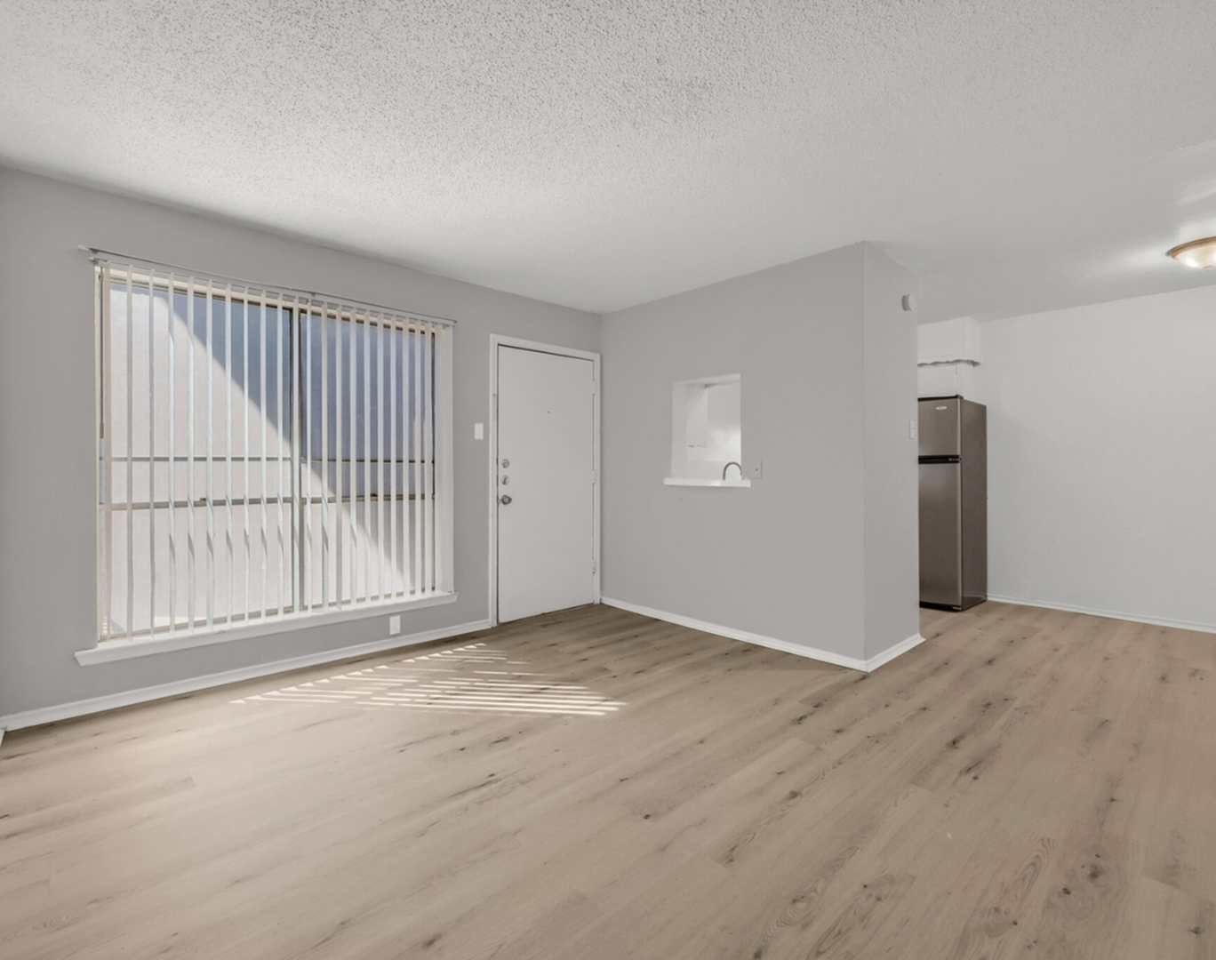 Bright and airy living room with large window and sleek wood flooring, adjacent to modern kitchen area.