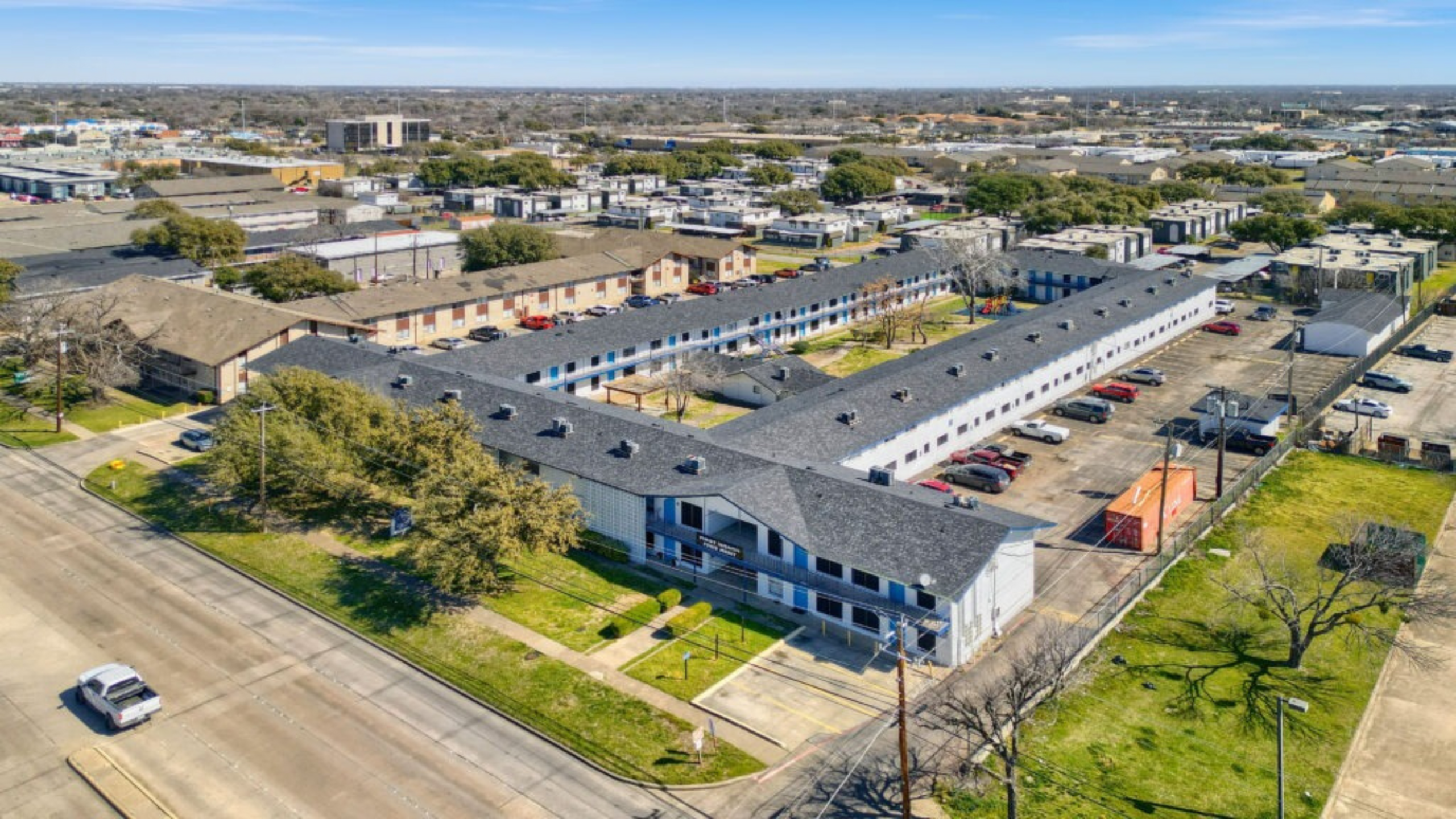 Aerial view of a modern apartment complex with stylish architecture and ample parking, surrounded by lush greenery and situated in a vibrant urban area.