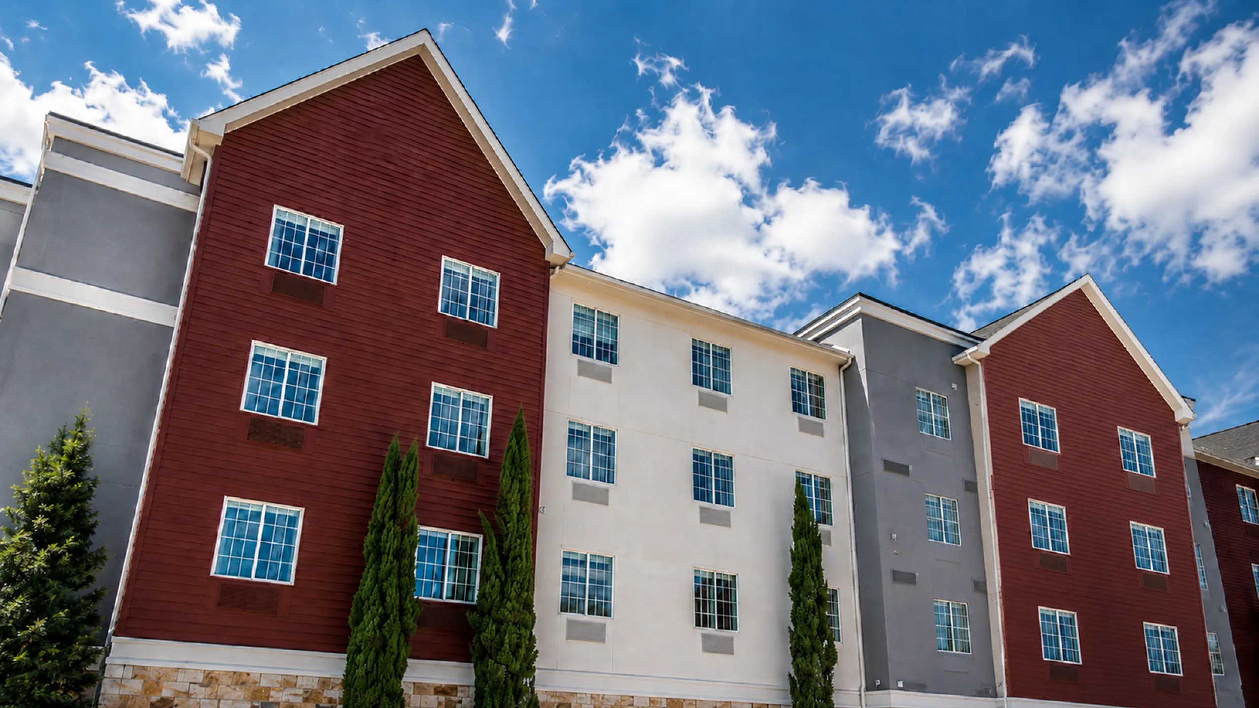 Charming apartment complex with brick and stucco facade, featuring large windows and beautiful landscaping against a bright blue sky.