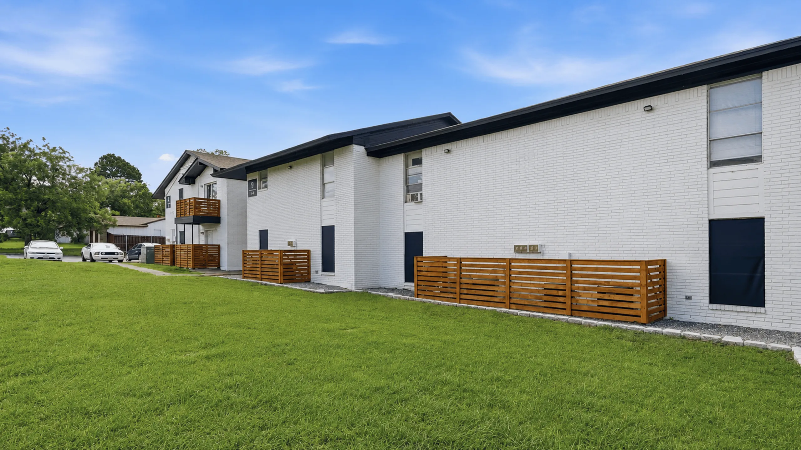 Modern apartment building with clean white brick exterior and charming wooden balcony accents surrounded by lush green lawn under clear blue skies.