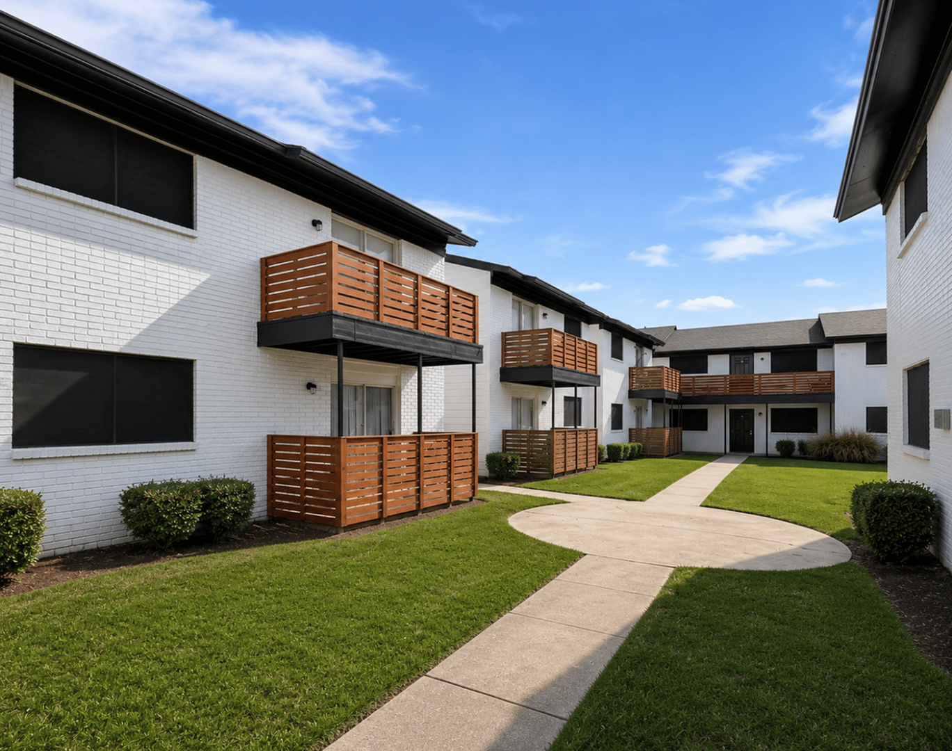 Charming apartment complex with modern wooden balconies, surrounded by lush green lawns and a clear blue sky backdrop.