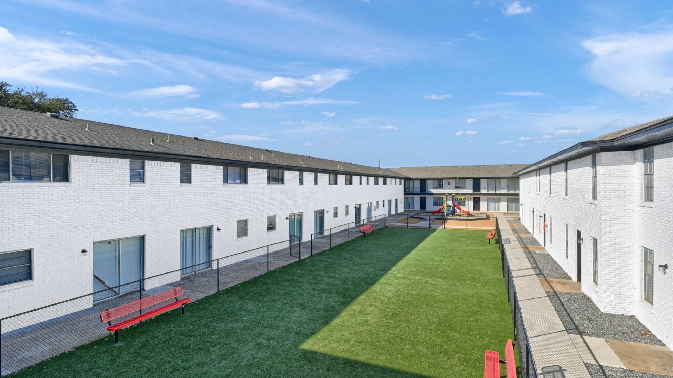 Inviting apartment courtyard with lush green space, vibrant red benches, and a modern playground under a bright blue sky.