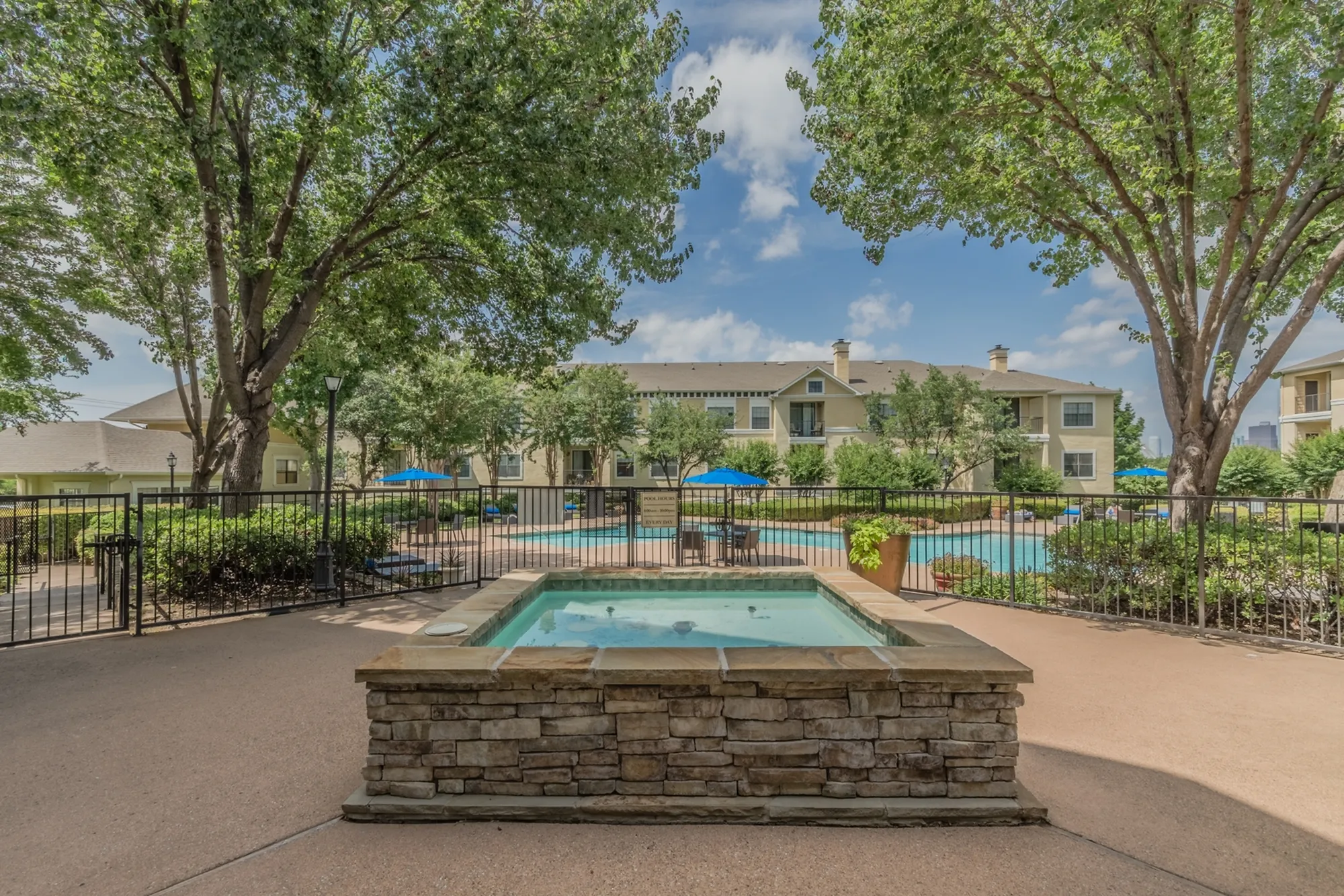 Luxurious apartment complex with a sparkling pool surrounded by lush greenery and elegant stone landscaping under a bright blue sky.
