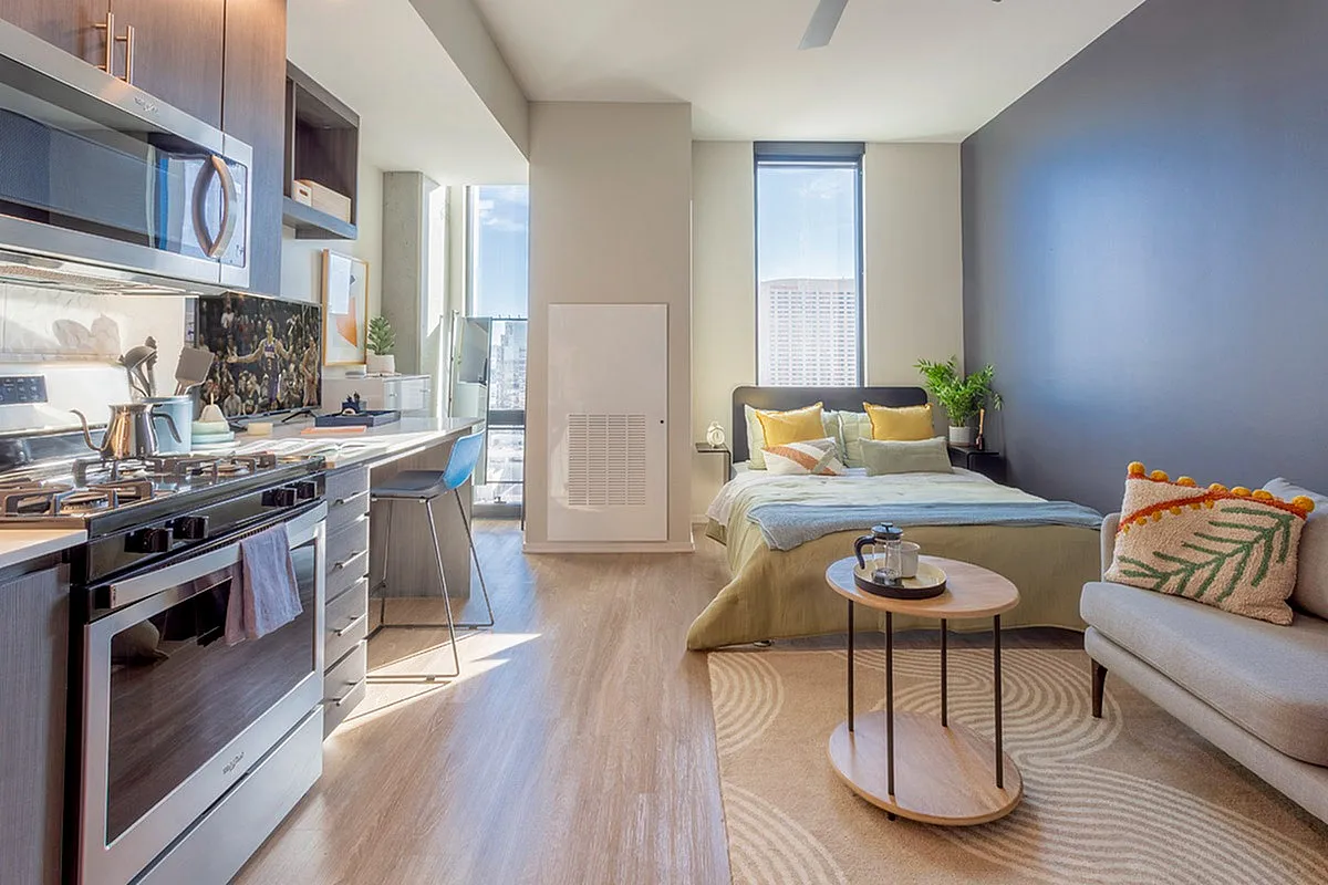 Bright and modern studio apartment featuring a cozy bed, sleek kitchen with stainless steel appliances, and a stylish seating area with natural light streaming through large windows.