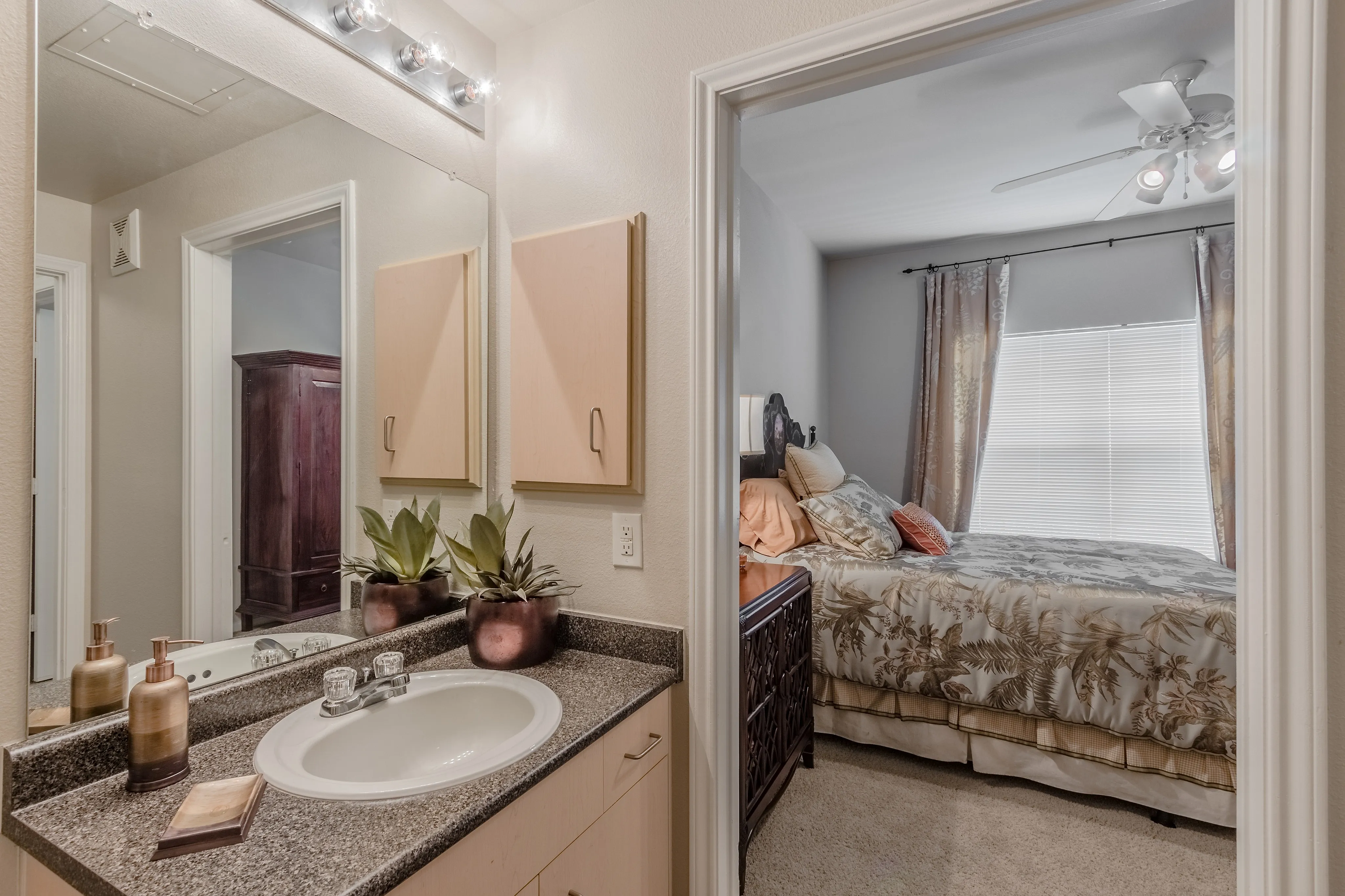Modern apartment bathroom and bedroom view with elegant decor, featuring a granite countertop, stylish sink, lush potted plants, and a cozy, well-lit bedroom with chic curtains and a comfortable bed.