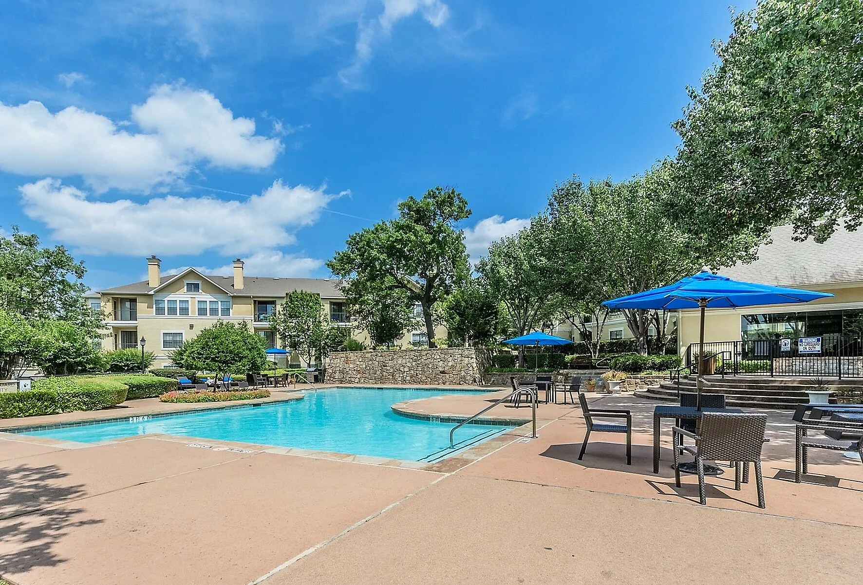 Inviting apartment complex with a sparkling pool, vibrant blue parasols, and lush greenery under a clear blue sky.