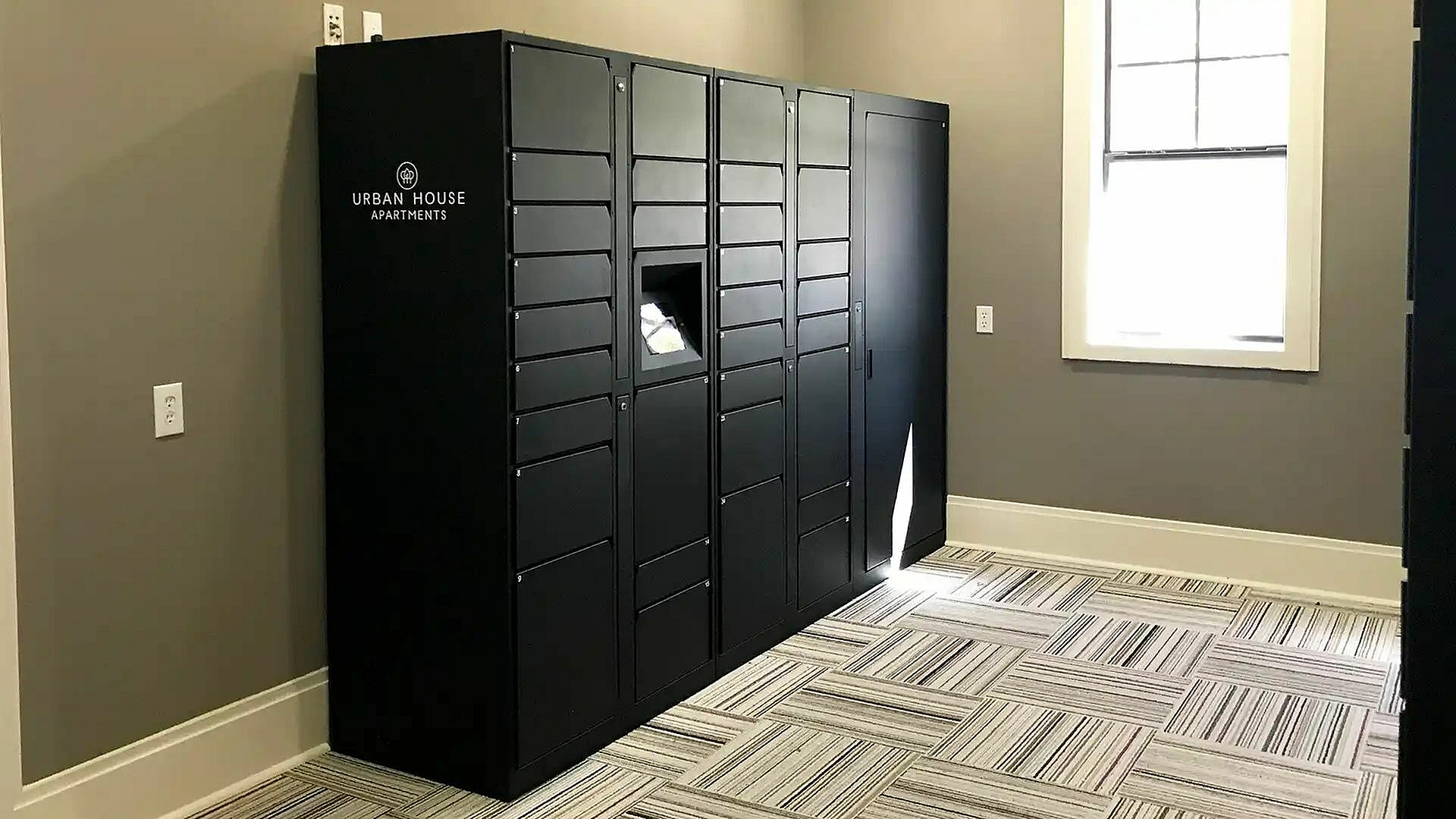 Modern package locker room with sleek black lockers, striped carpet, and natural light from a large window, ideal for convenient urban living.