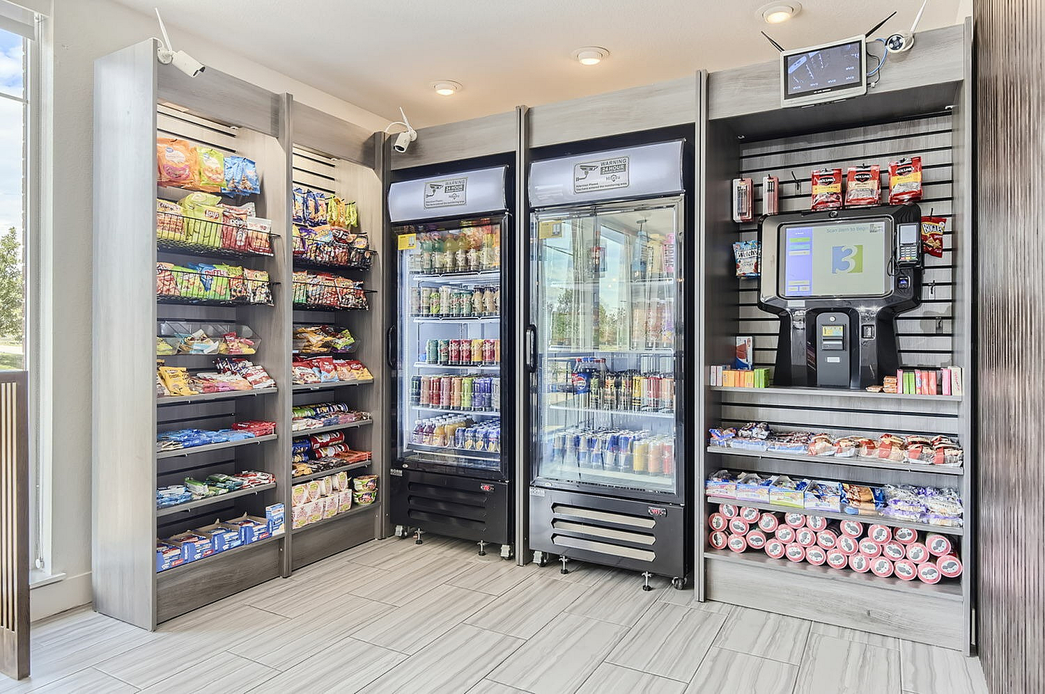 Modern apartment convenience store with stocked snack shelves, refrigerated beverages, and a self-checkout station, offering a bright and welcoming shopping experience.