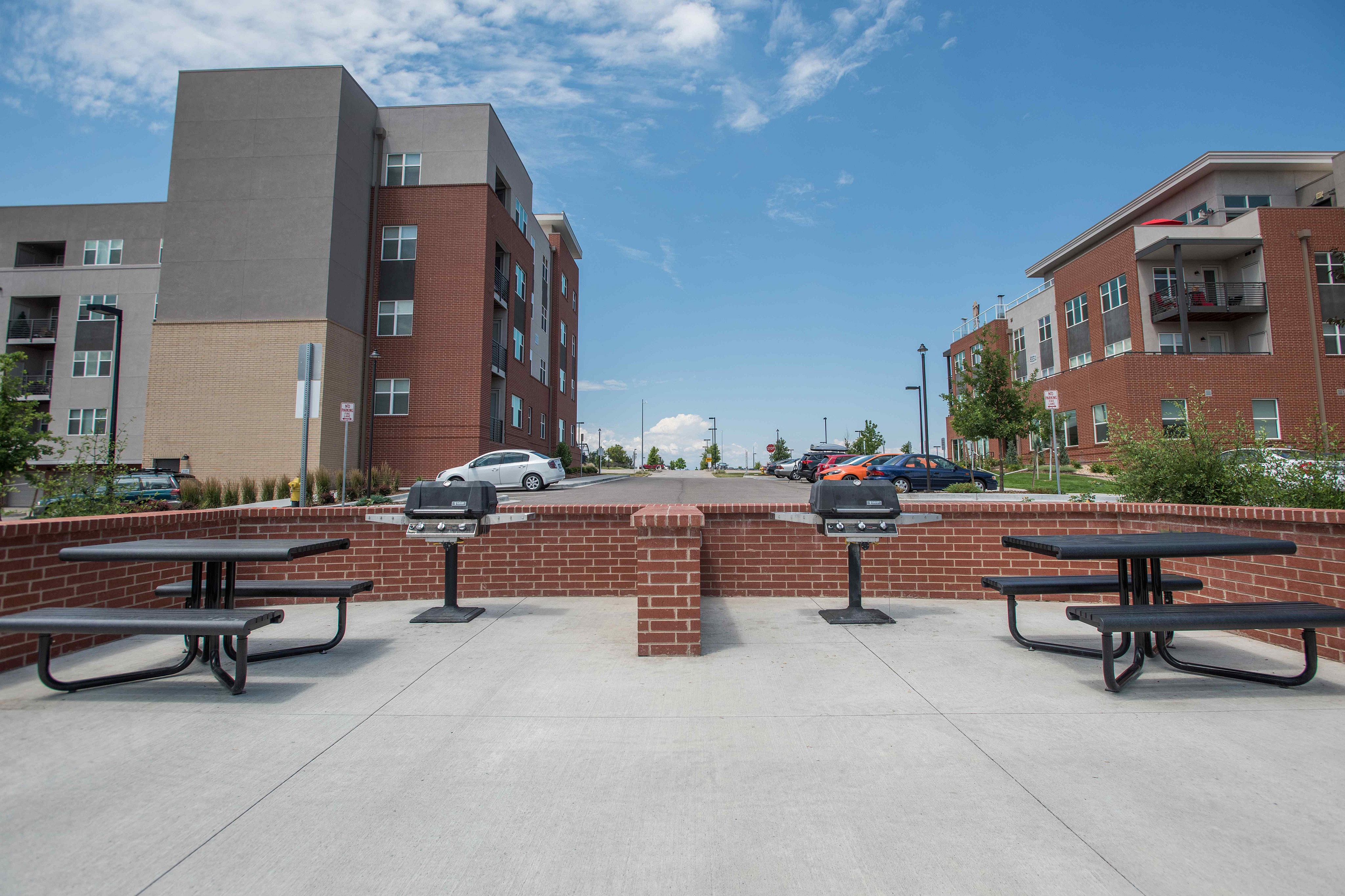 Modern apartment complex featuring stylish brick architecture, ample parking, and outdoor amenities including picnic tables and grills, set under a bright blue sky.