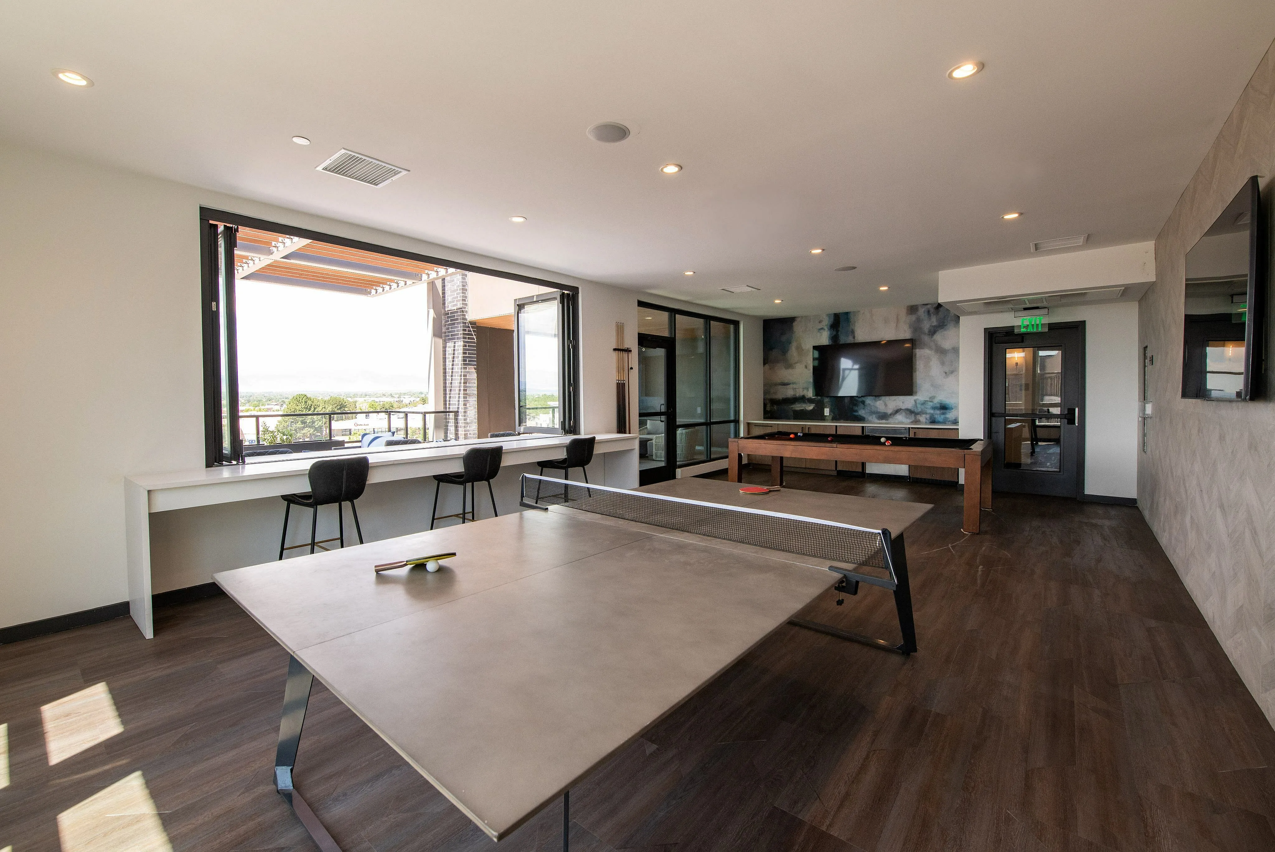 Bright recreational room with large windows, featuring a ping pong table, pool table, and outdoor view seating.