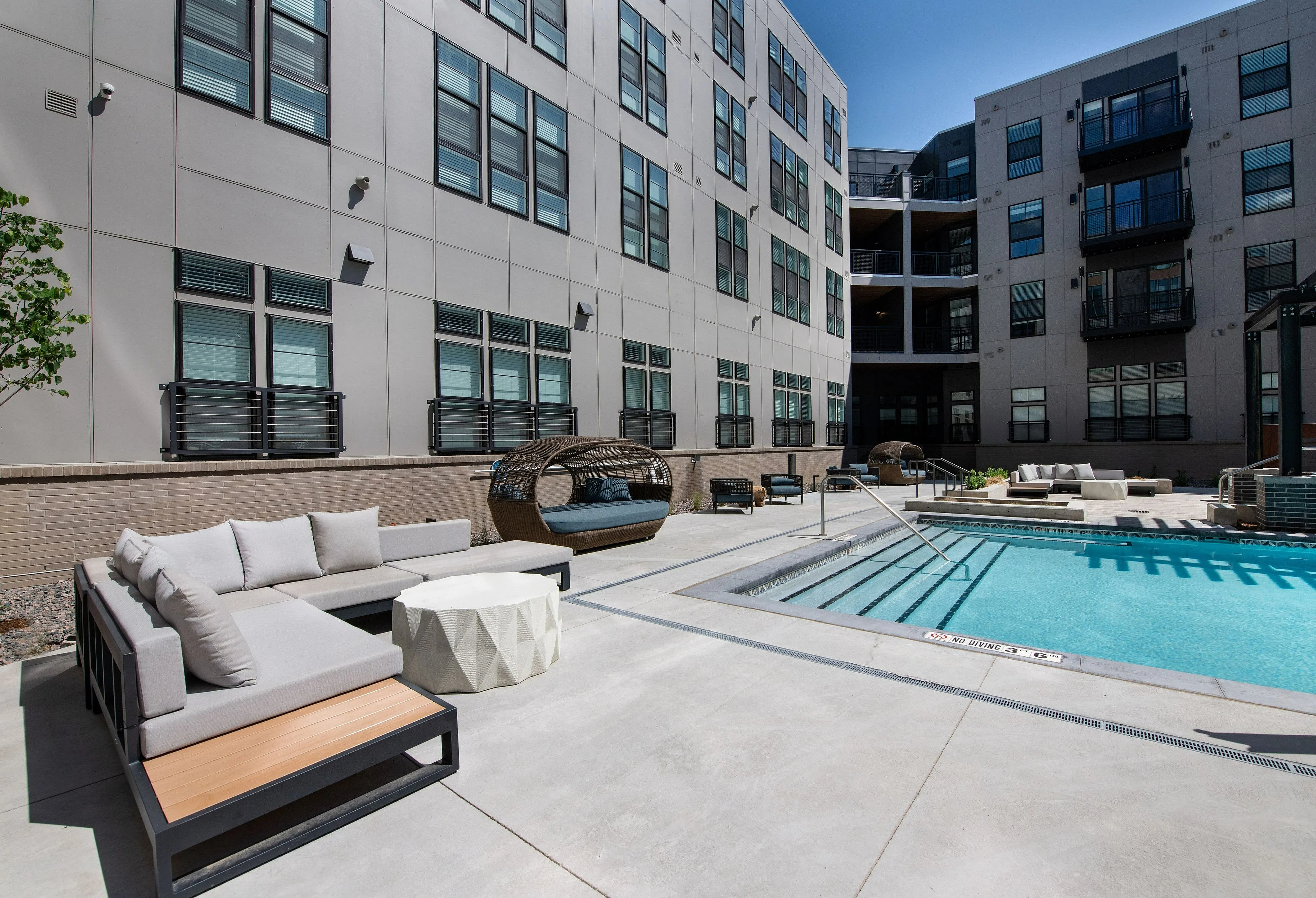 Modern apartment building with chic outdoor patio featuring stylish seating, inviting pool, and relaxing sunbeds under a clear sky.