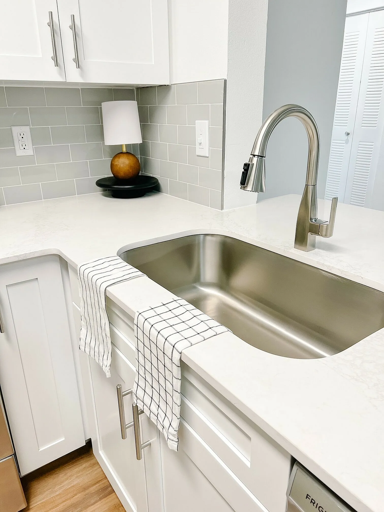 Modern kitchen corner with sleek stainless steel faucet, elegant white cabinetry, and chic subway tile backsplash, accented by a stylish table lamp.