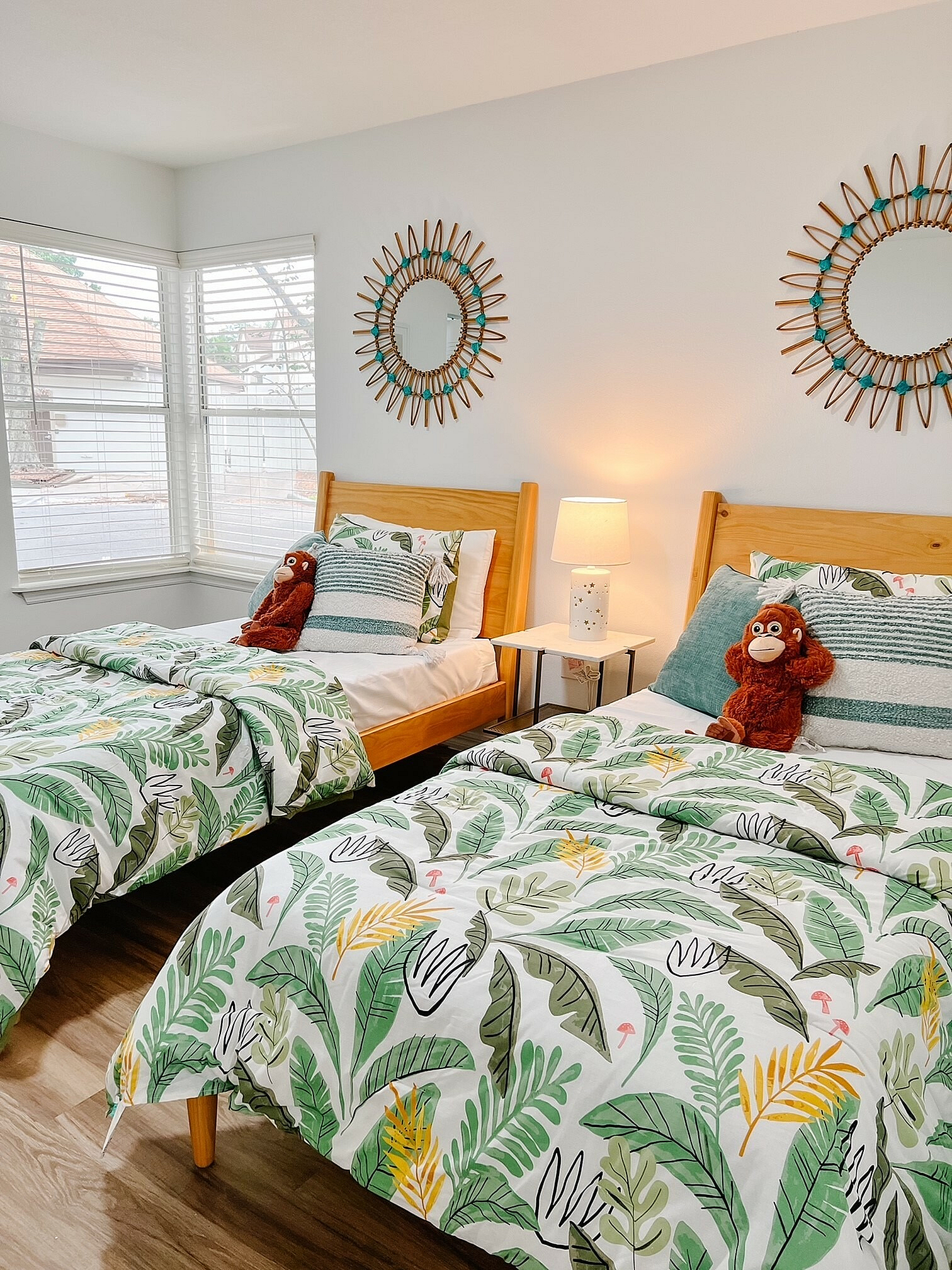 Bright and charming bedroom with twin beds, tropical-themed bedding, warm wooden furniture, and decorative round mirrors creating a cozy, inviting atmosphere.