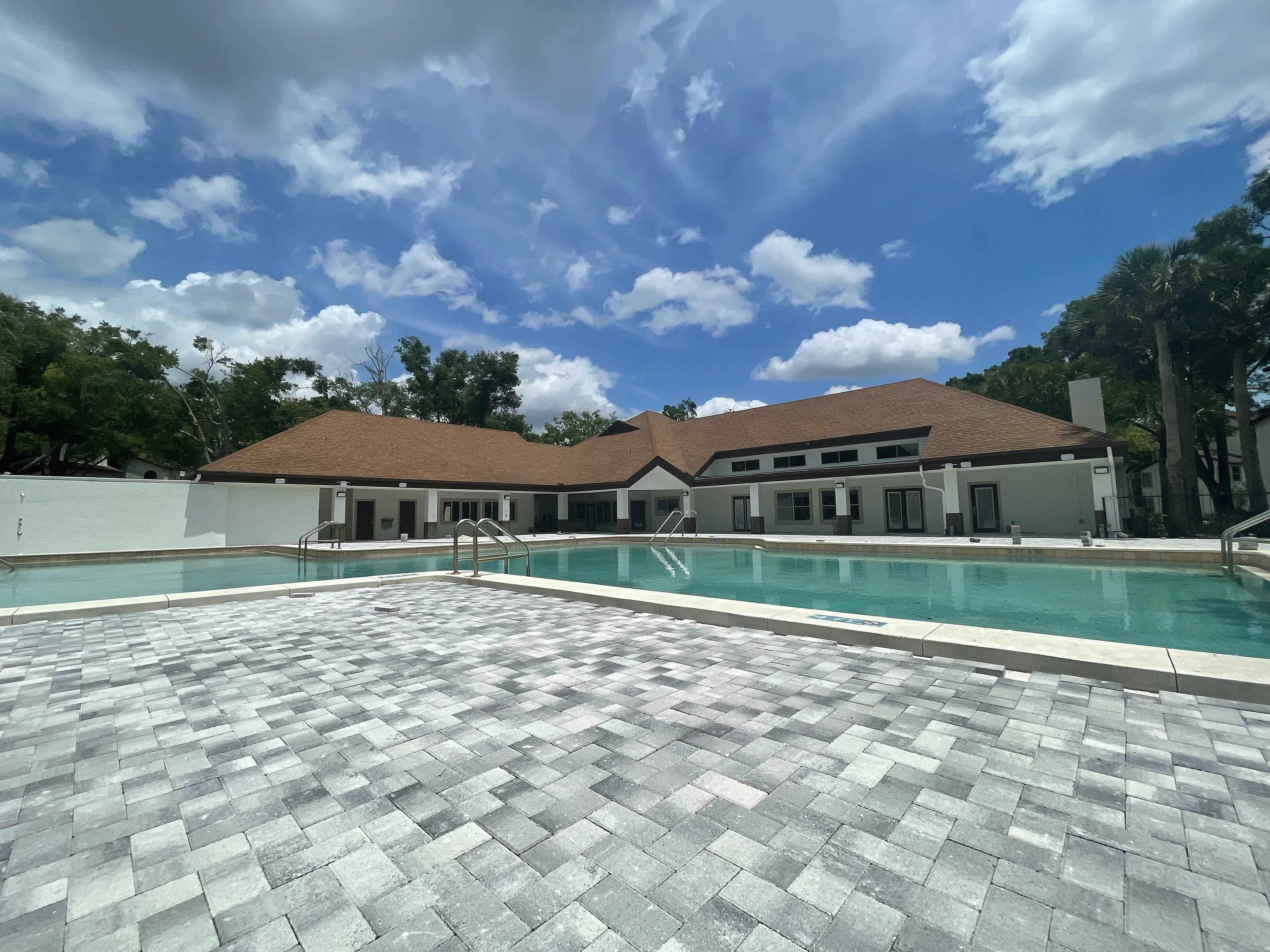 Inviting outdoor pool area with modern paver deck and spacious clubhouse under a bright blue sky.