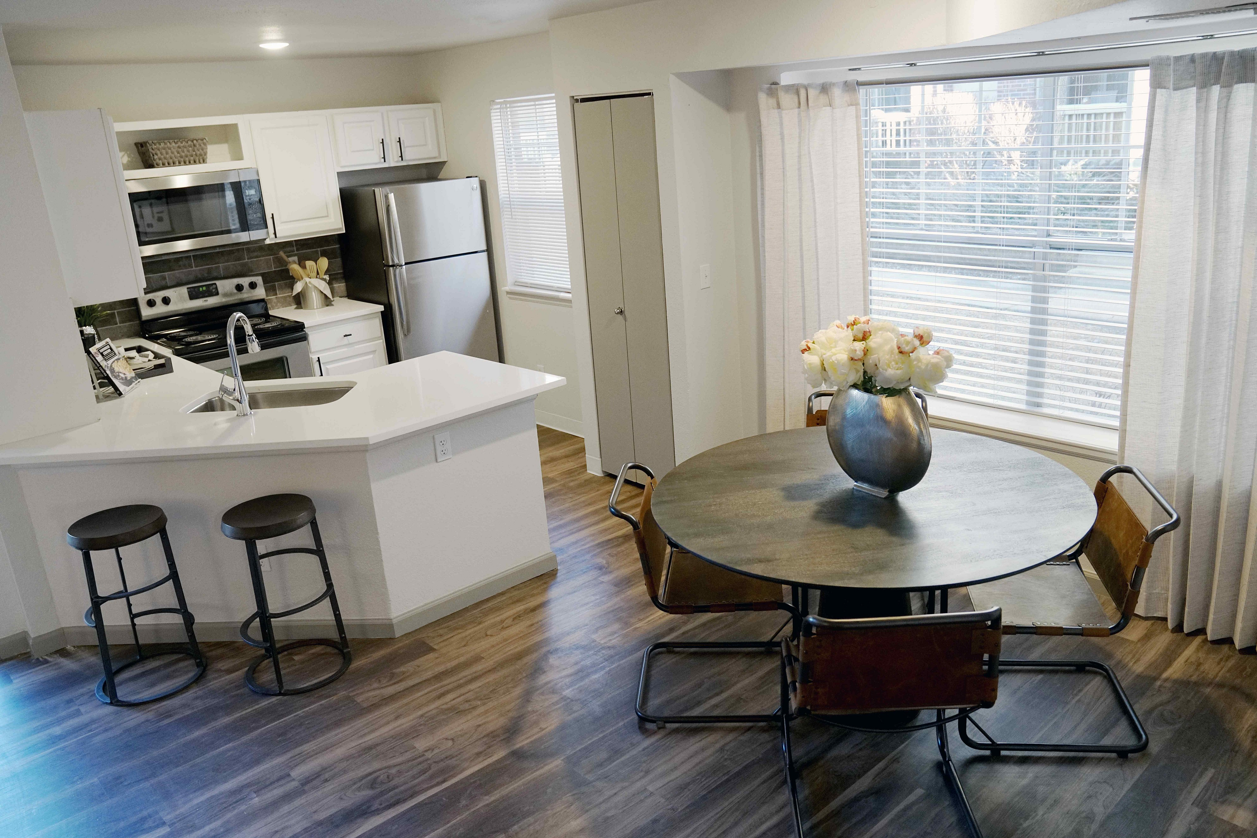 Chic kitchen and dining area featuring stainless steel appliances, modern white cabinetry, and cozy breakfast bar with stools. Bright dining nook with round table and elegant flower centerpiece.
