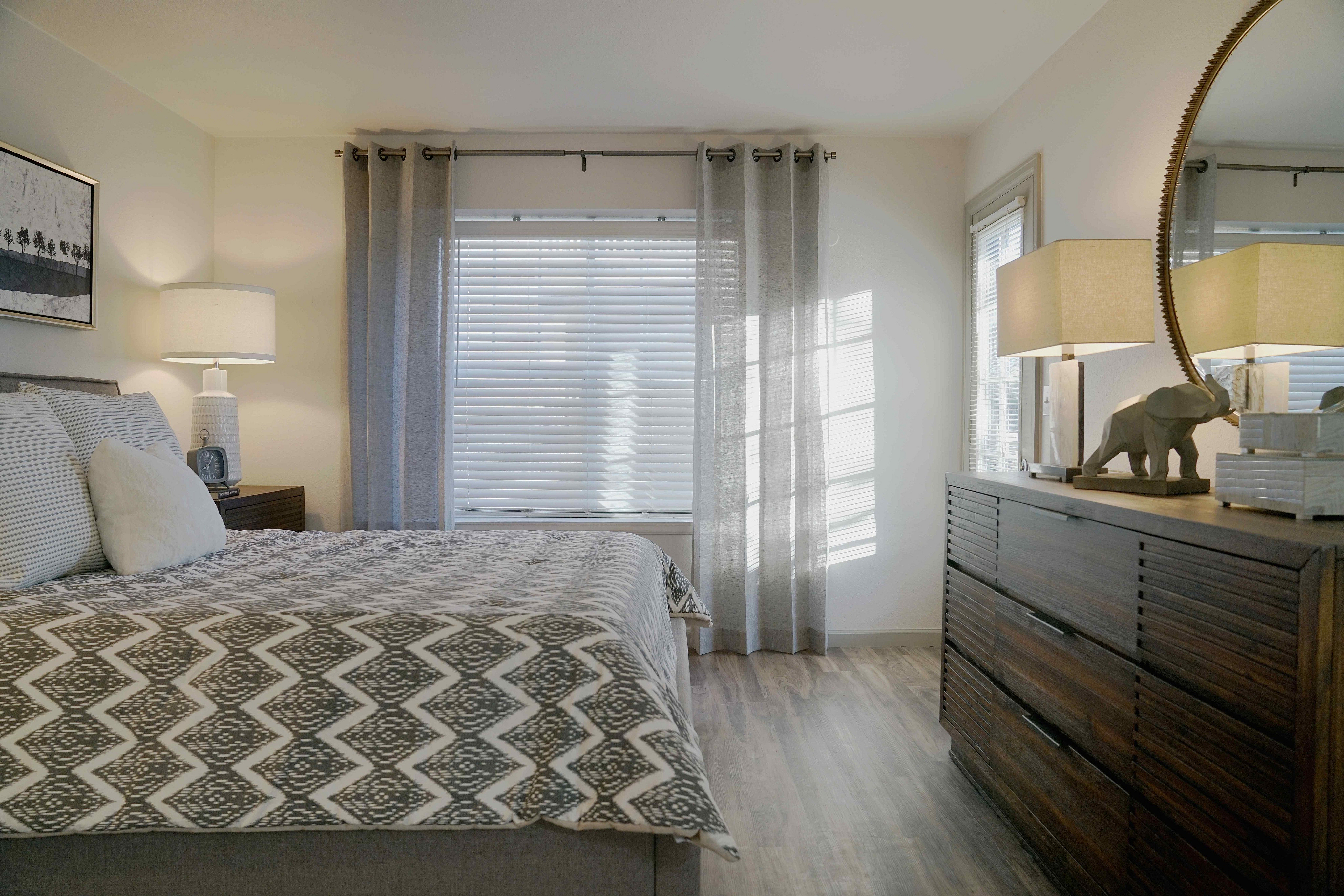 Inviting bedroom with abundant natural light, featuring elegant decor, plush bedding, and a stylish dresser accented by a decorative lamp.