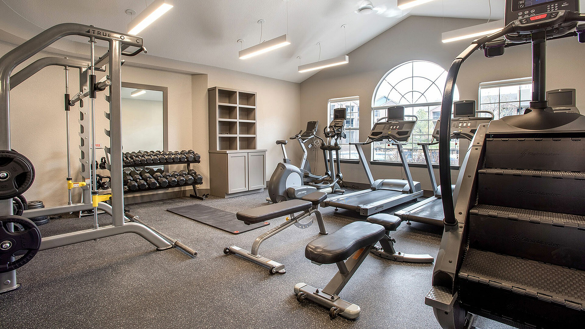 Modern fitness center with state-of-the-art gym equipment, including treadmills, weight machines, and a StairMaster, illuminated by natural light through large arched windows.