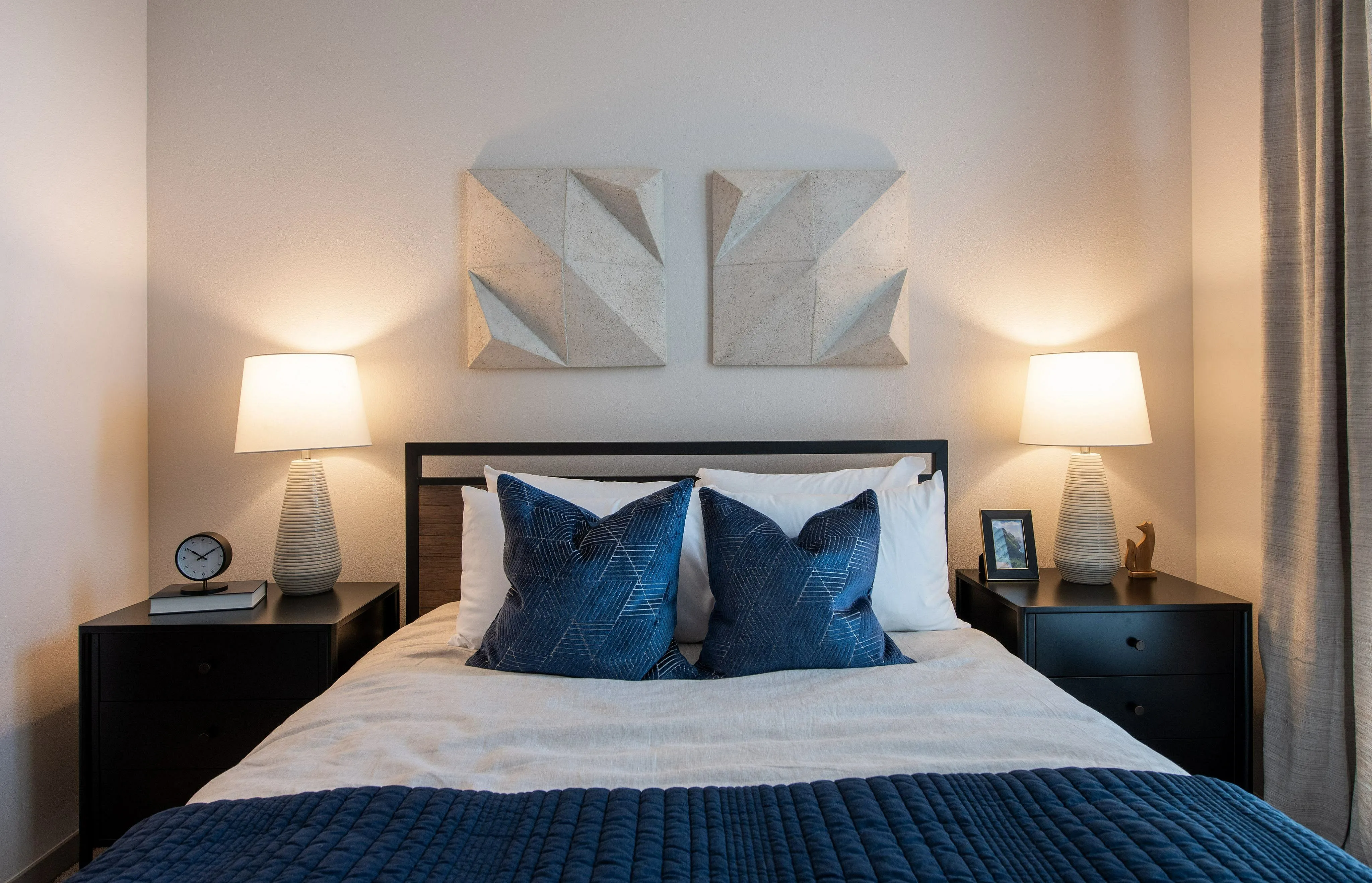 Elegant bedroom with modern decor, featuring a cozy bed adorned with blue cushions, stylish geometric wall art, and soft ambient lighting from bedside lamps.
